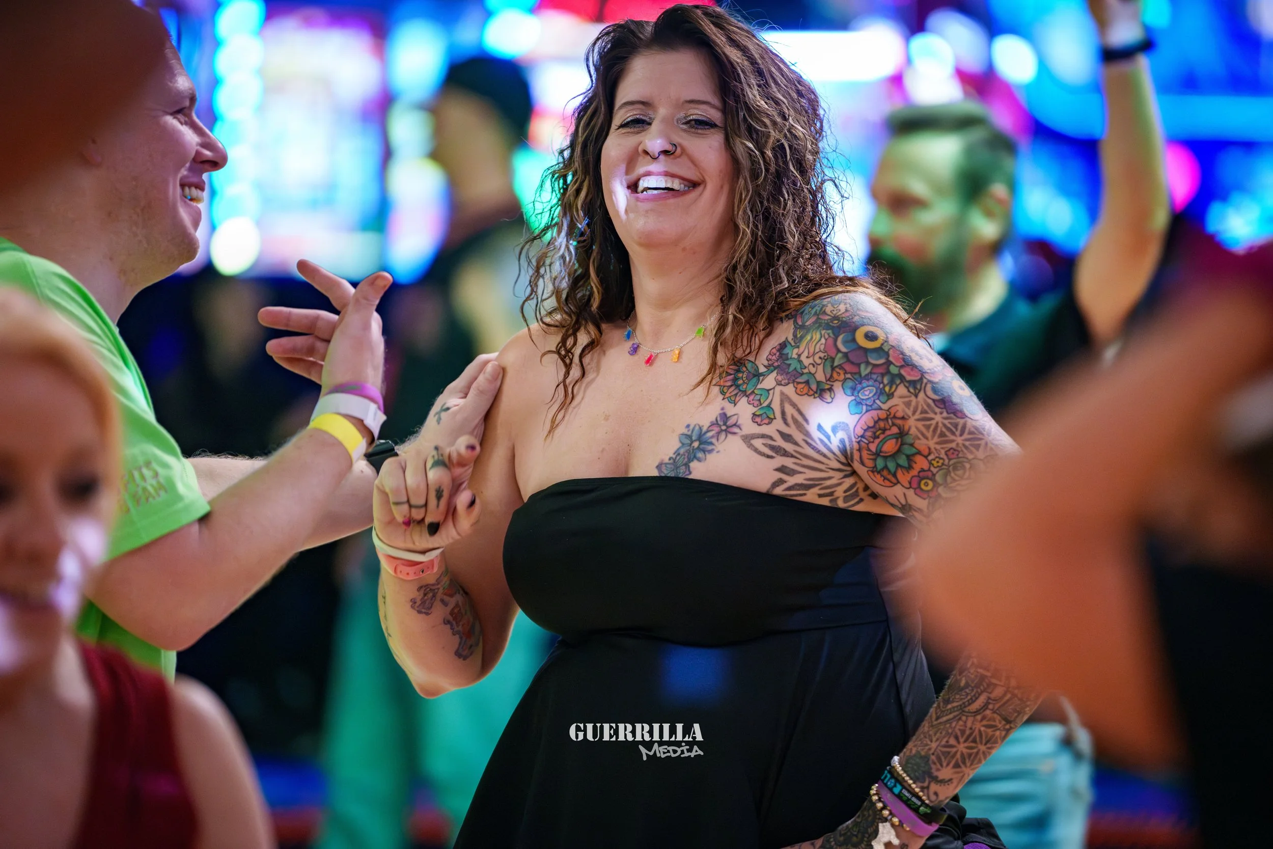 A woman with curly hair and tattoos on her shoulder and arm, smiling and dancing at a lively event with colorful lights in the background.