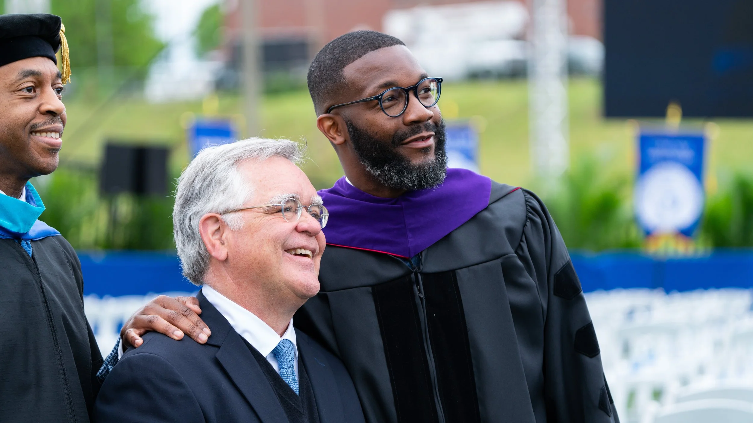 Three men at a graduation ceremony, one in graduation robes and cap, two in business or academic attire, all smiling, outdoors with a blurred background of greenery and banners.