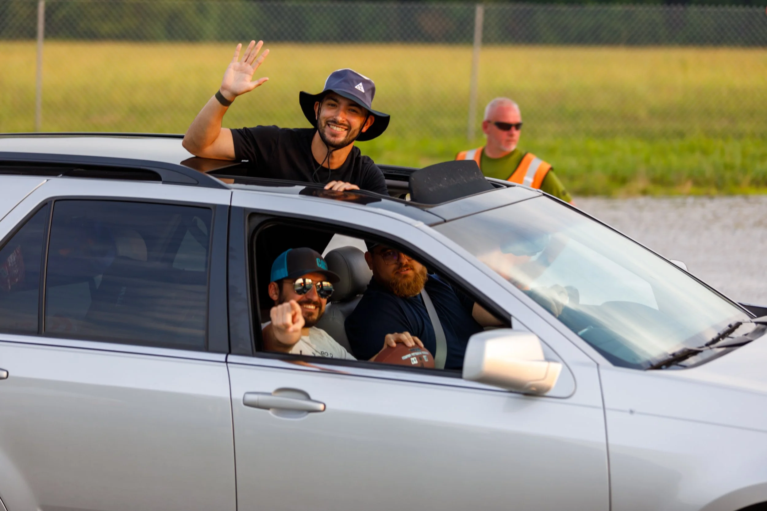 Four men inside a silver SUV at a daytime outdoor location. One man is leaning out of the sunroof, waving and smiling. The driver and passenger are both wearing sunglasses and looking forward. The man leaning out is wearing a black shirt and a wide-b