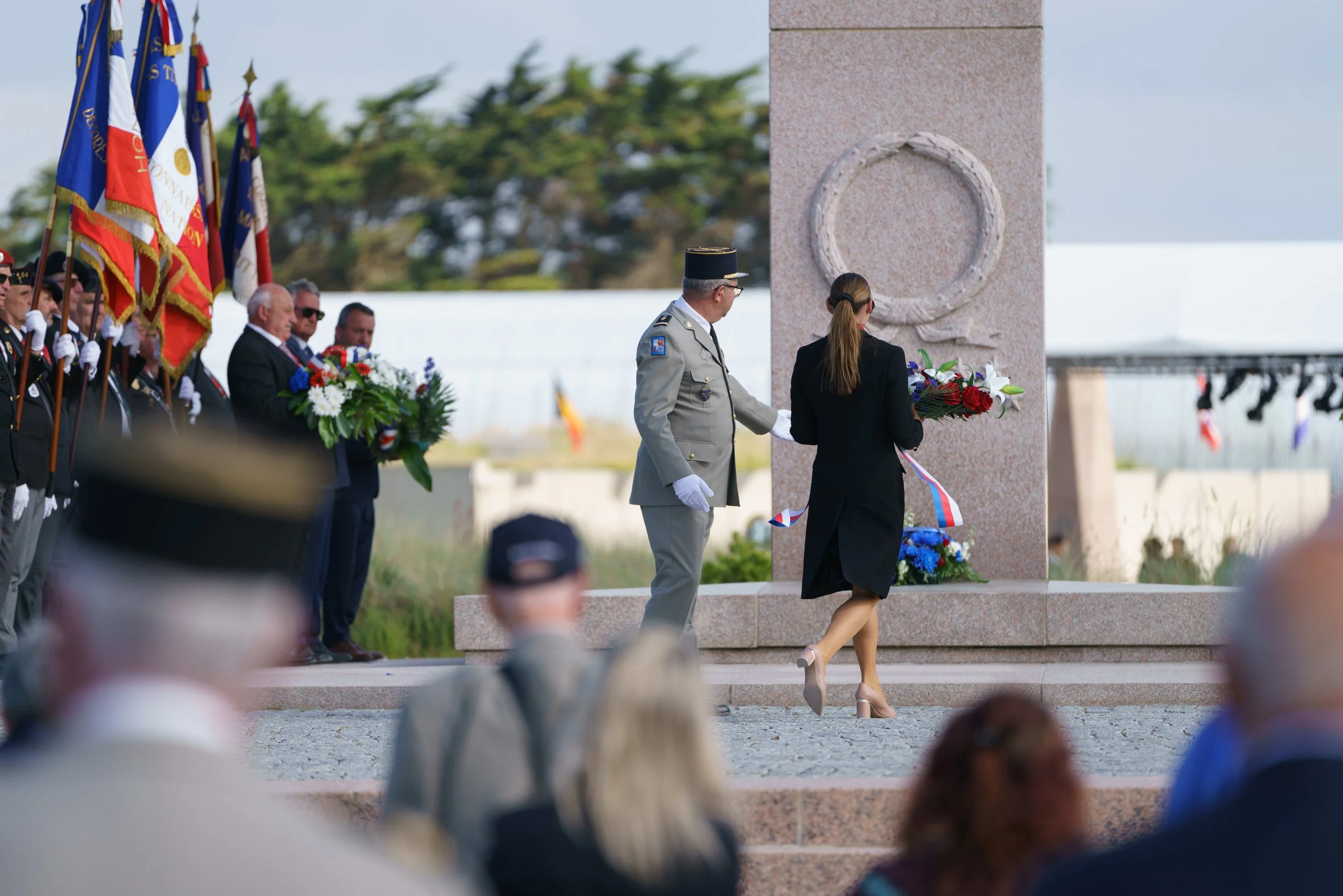 A memorial ceremony with individuals laying flowers at a monument, with a group of people holding flags and a crowd observing, outdoors during daylight.