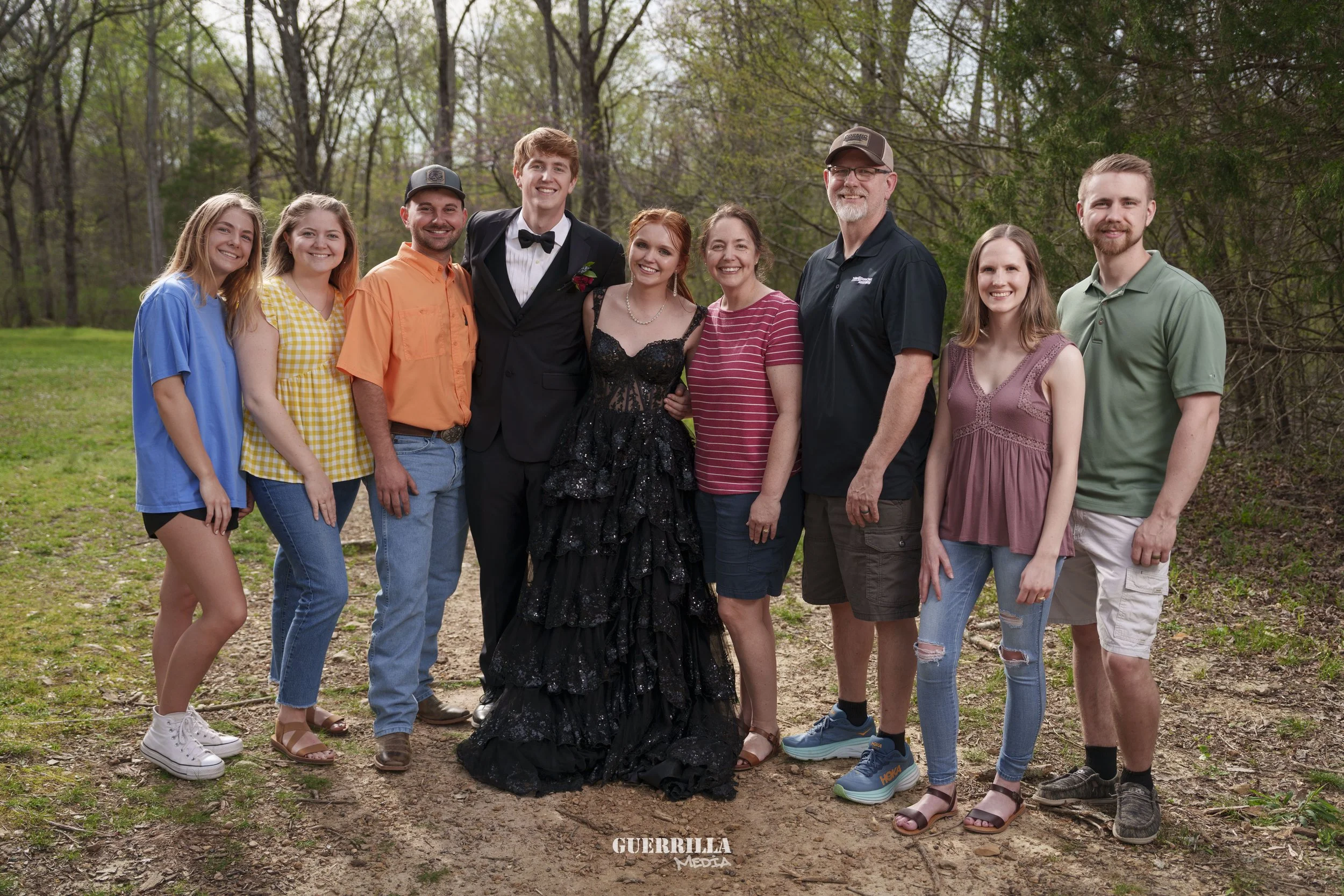 A group of nine people standing outdoors on a dirt path in a wooded area, dressed casually and semi-formally, smiling at the camera, with some wearing colorful summer clothes and one person in a black dress and tuxedo.