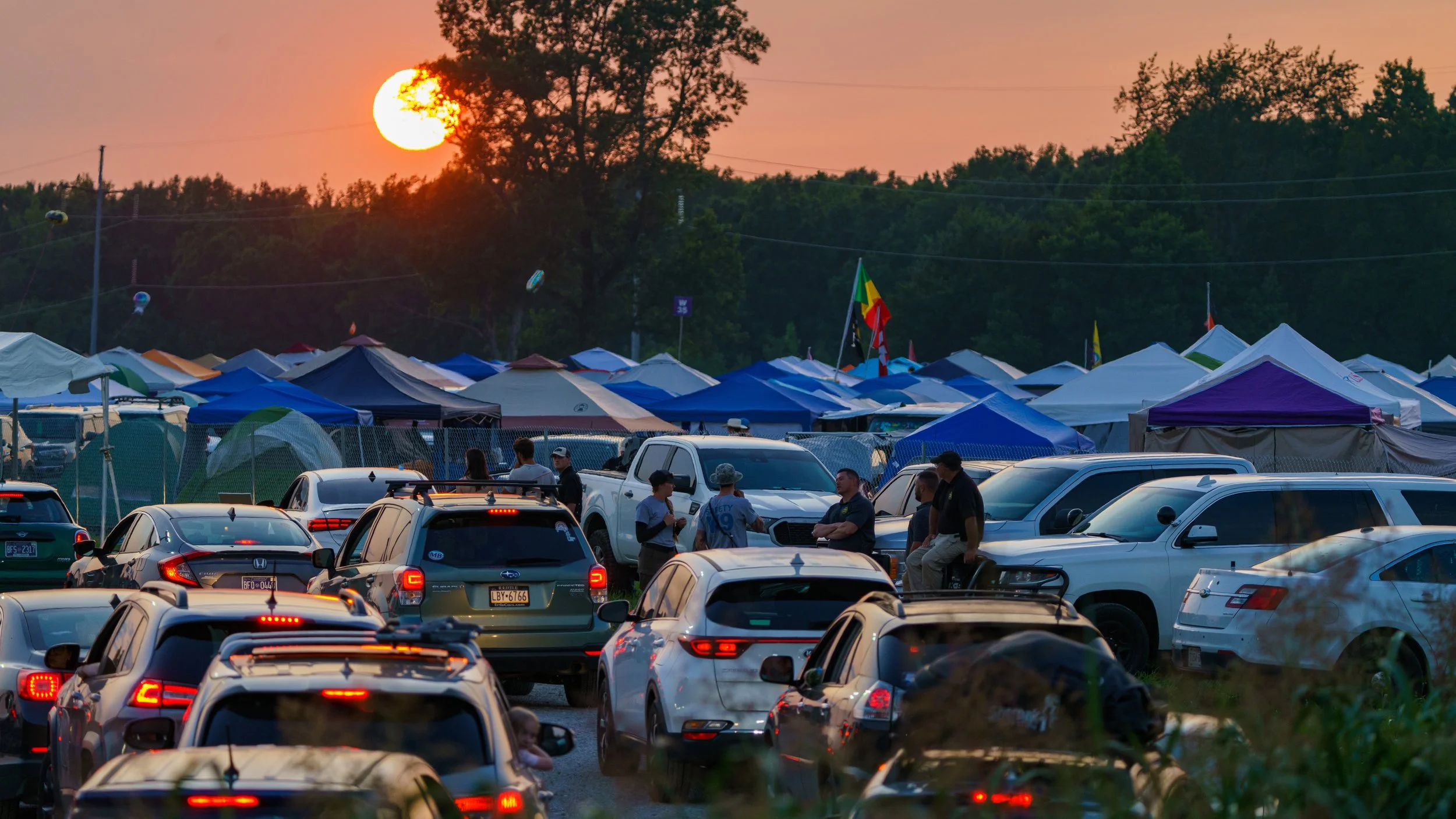 Sunset over a campsite and parking lot filled with cars and tents, with a group of people chatting near the parked vehicles and trees in the background.