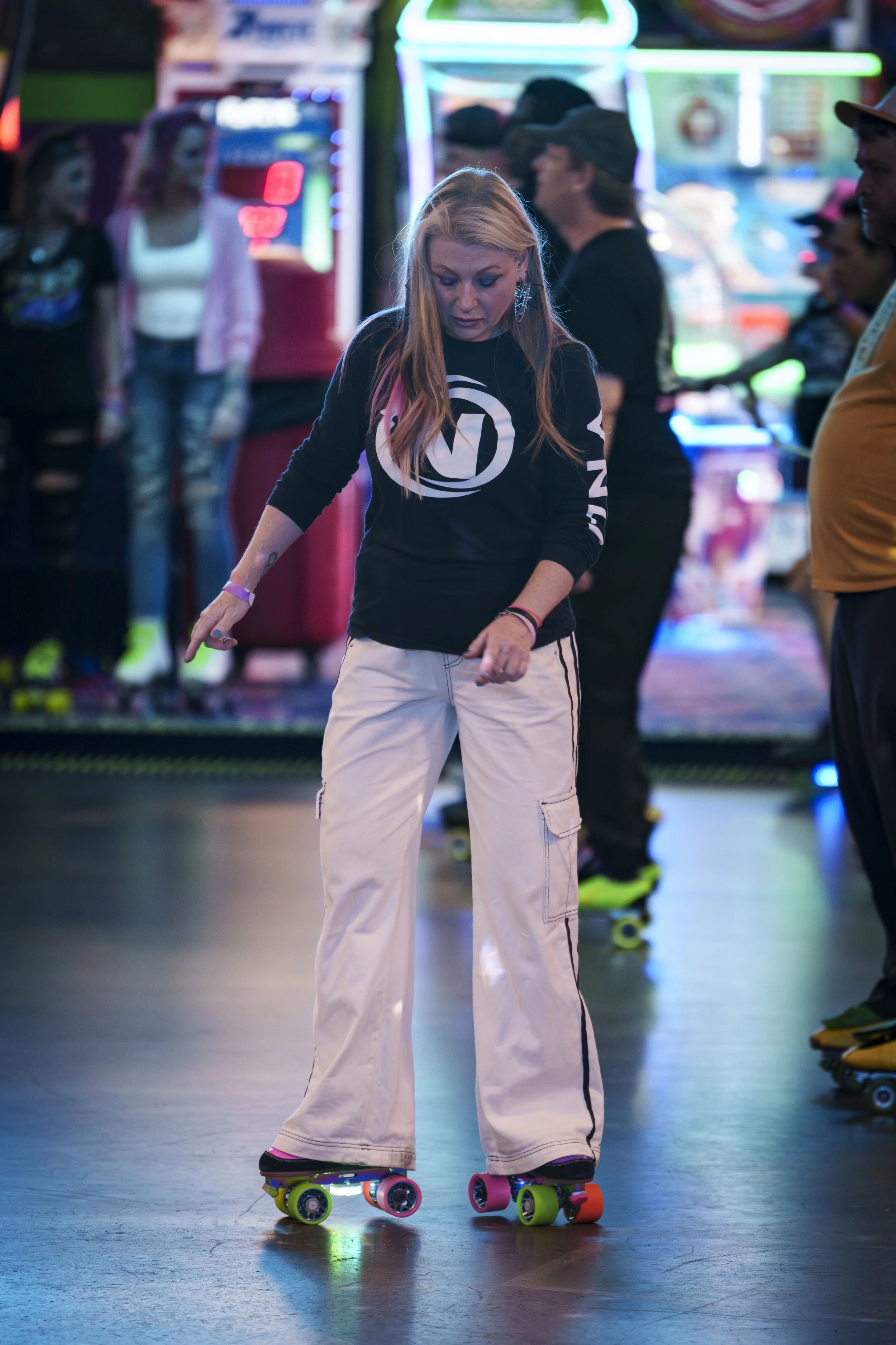 A young woman roller skating in an indoor arcade with neon lights, wearing a black shirt with a white logo and beige cargo pants.