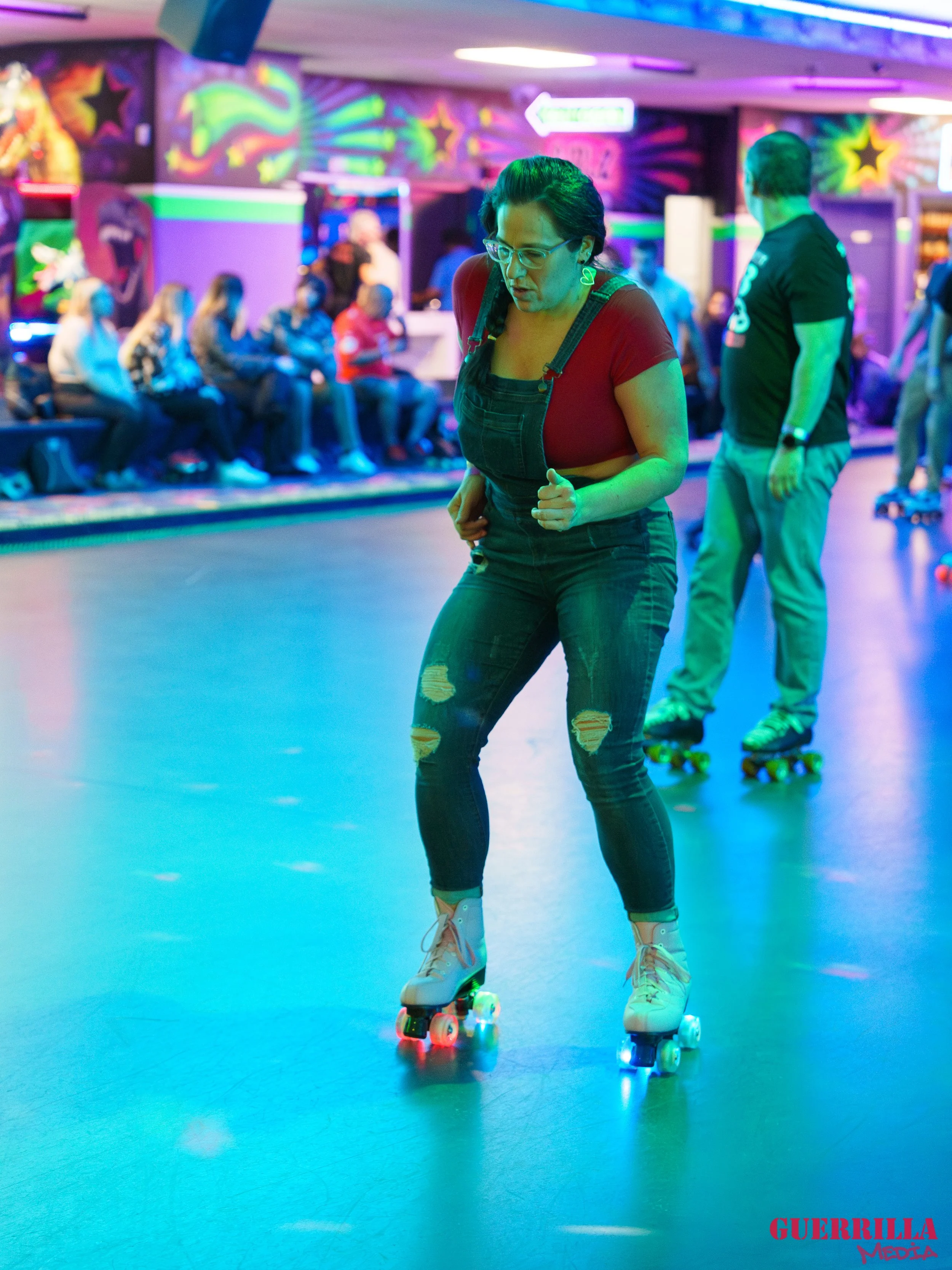 A woman roller skating in an indoor skating rink with colorful neon lights, wearing a red shirt, ripped jeans, and glasses, with a group of people seated in the background.
