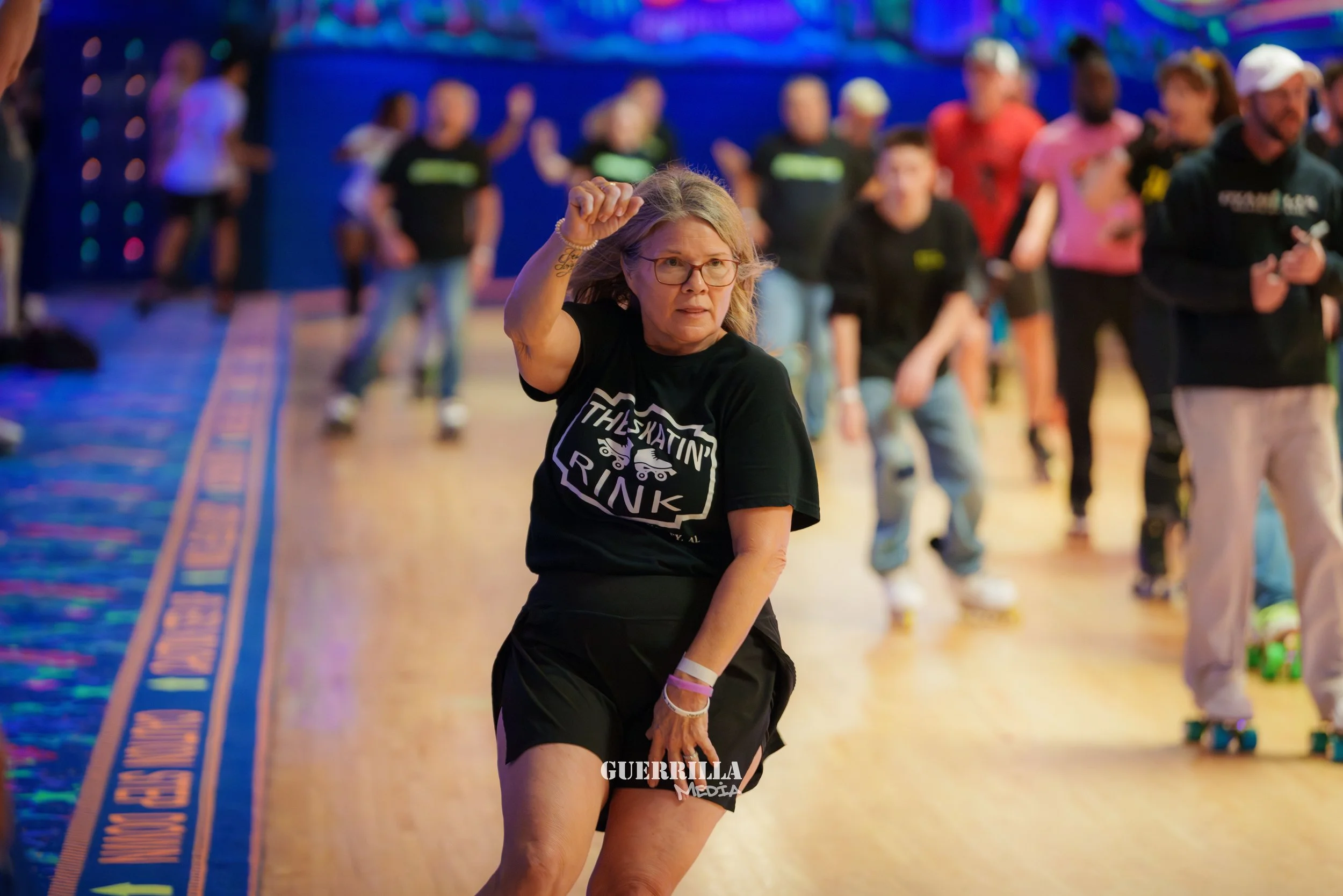 An elderly woman roller skating indoors with a group of people in the background, some wearing pink and black shirts, in a venue with colorful lighting.
