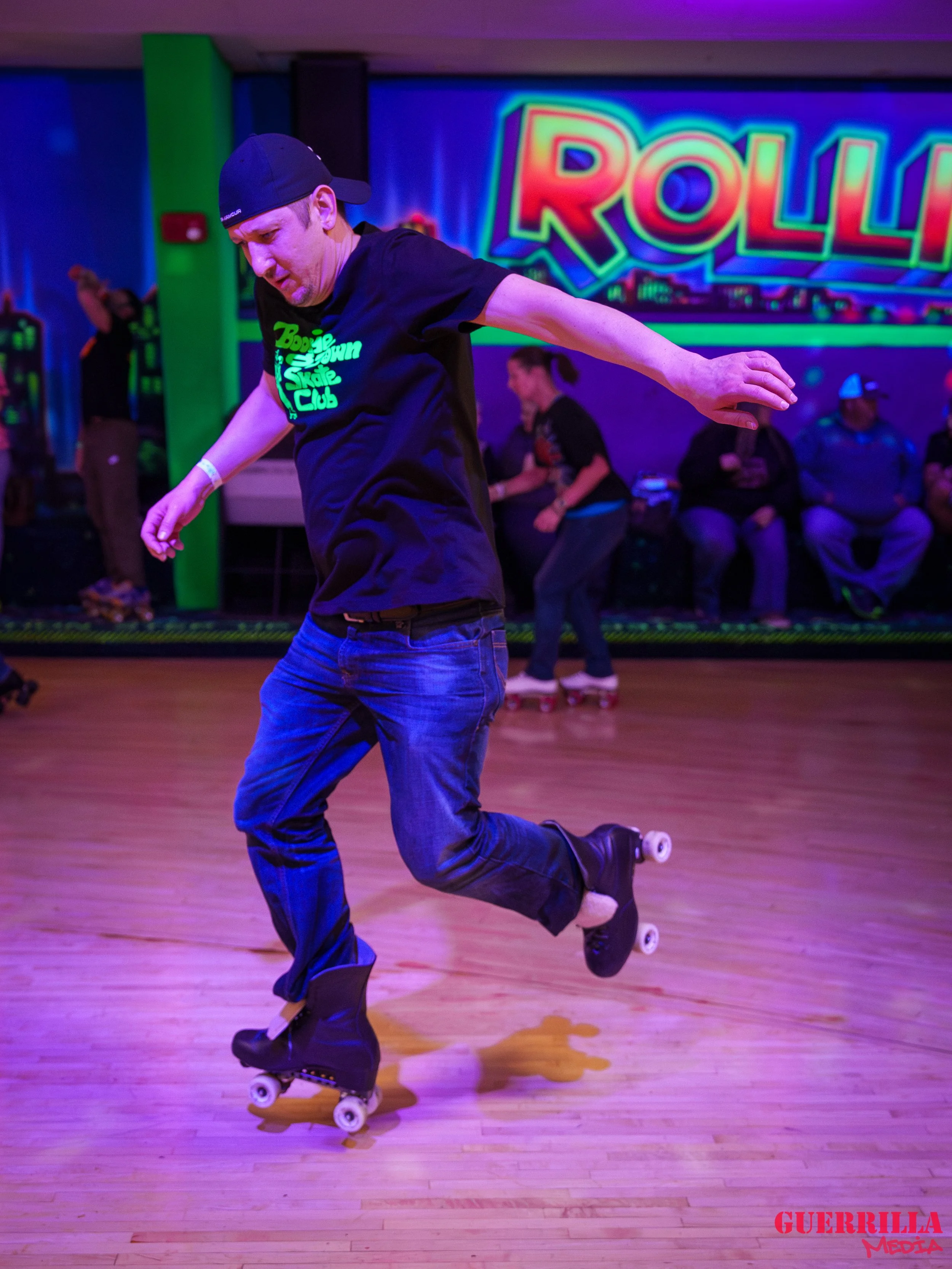 A person roller skating indoors with a neon-lit sign that says 'ROLLA' in the background. The individual is balancing on one leg with arms extended, in a skate rink setting.