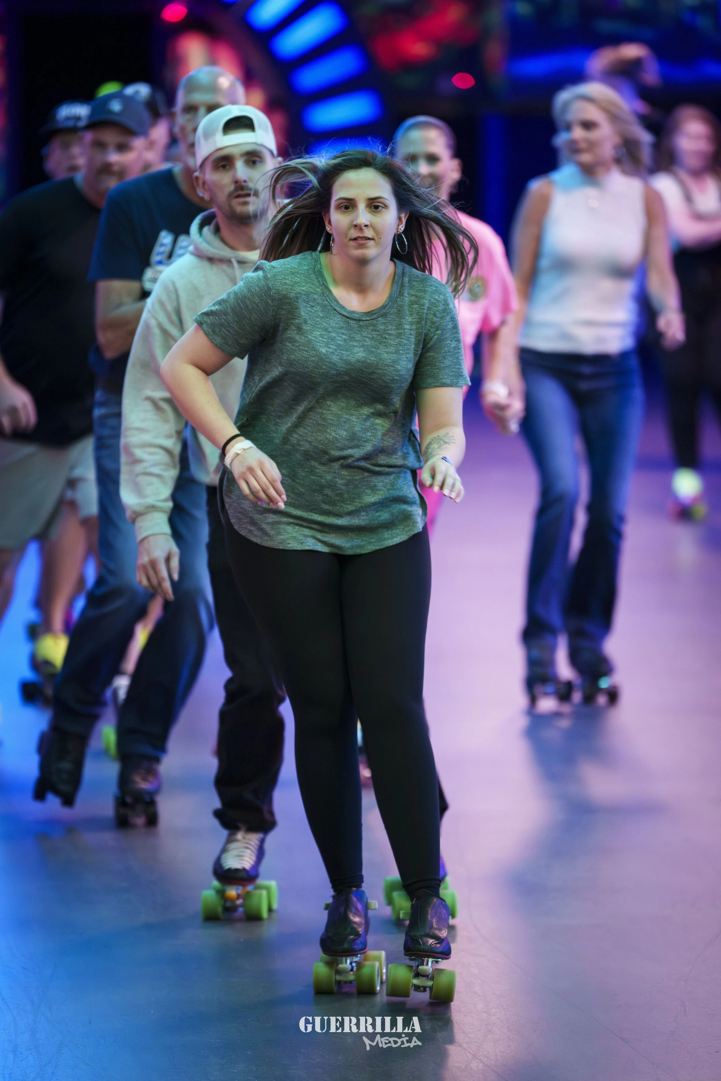 Group of people roller skating indoors with colorful lighting, leading with a woman in a green t-shirt and black pants.