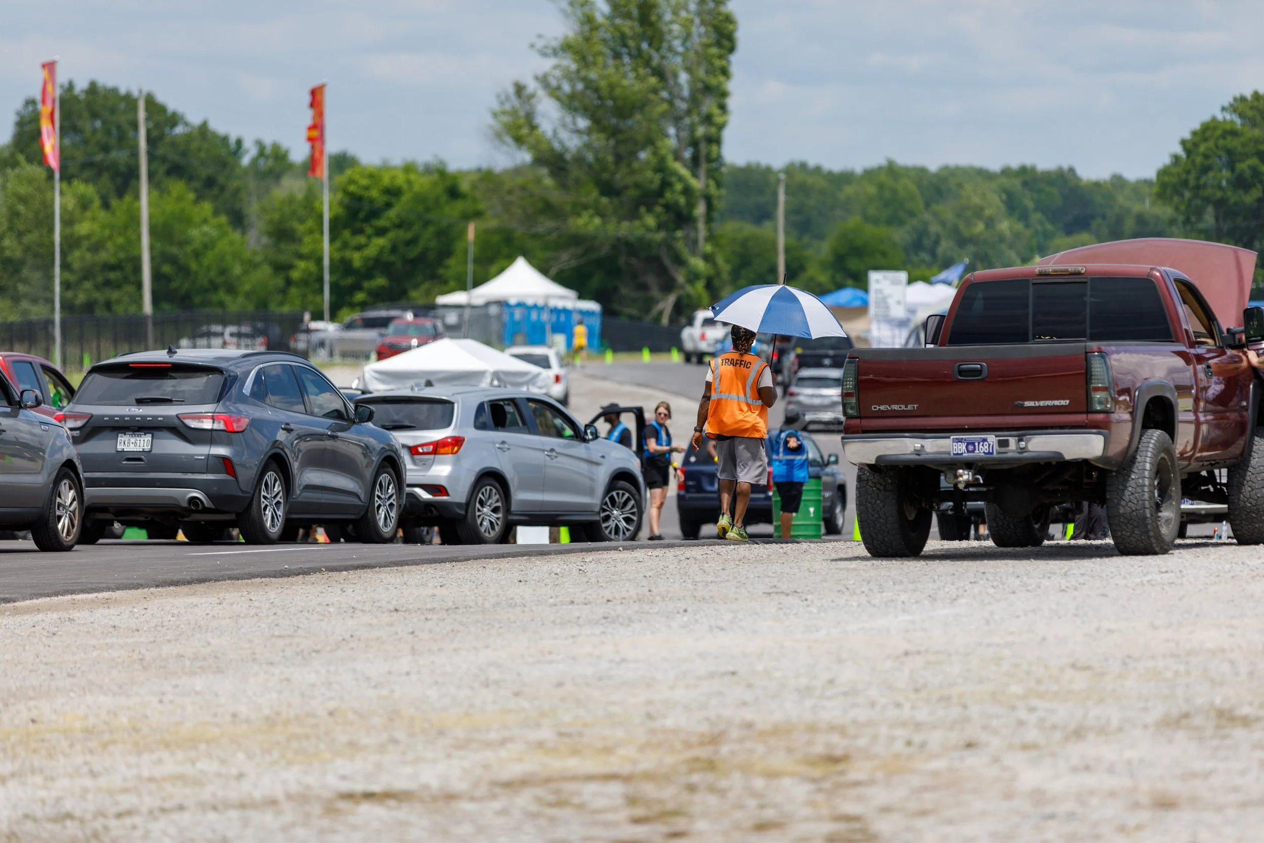 People at a parking lot during an outdoor event, with a traffic controller holding an umbrella, several cars including a red truck, and portable toilets in the background.