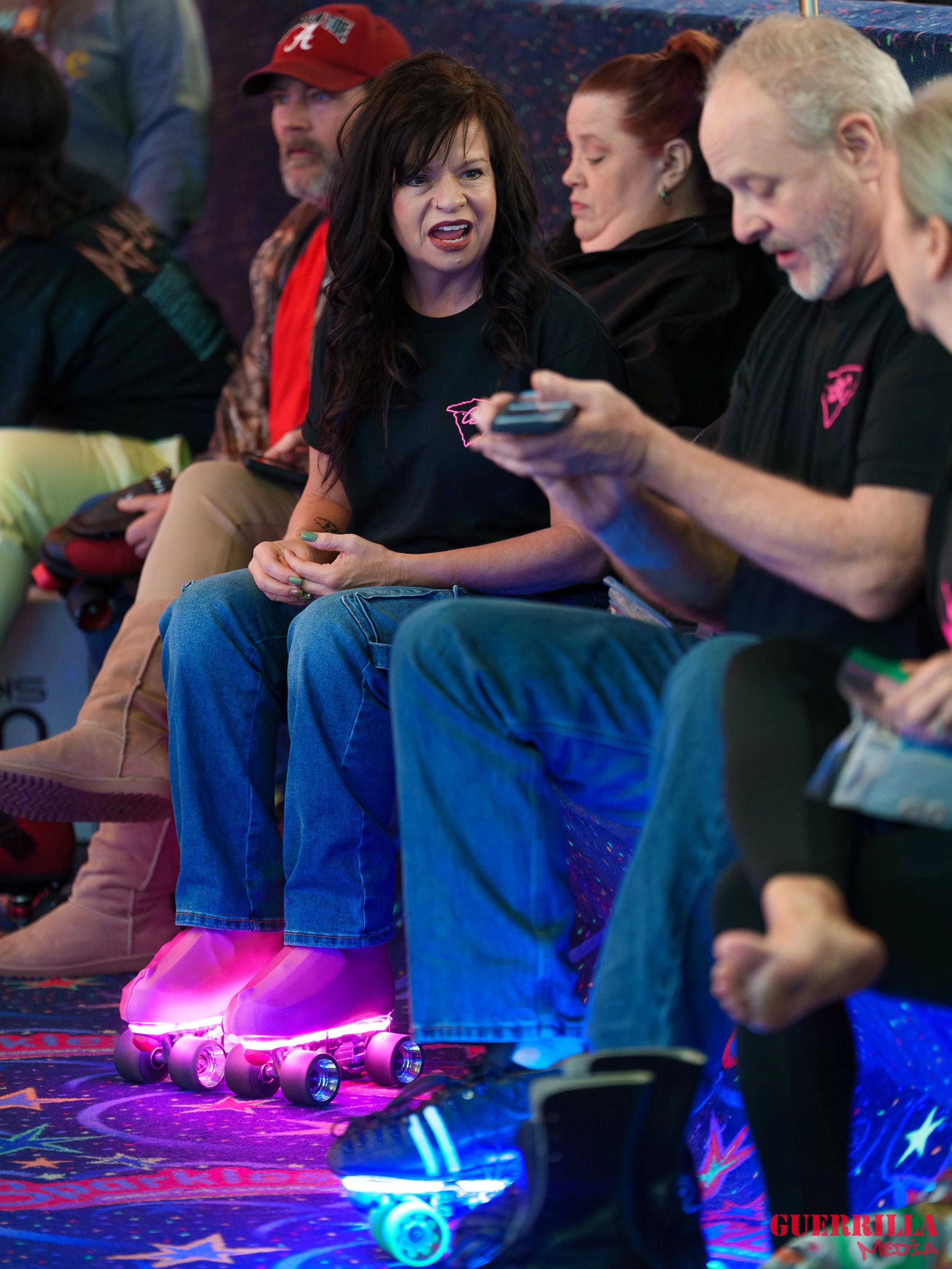 A woman with dark hair wearing a black t-shirt and blue jeans sitting on a bench, wearing pink roller skates with lights, and engaged in a conversation with a man holding a smartphone. Several other people are sitting nearby, some looking at their ph