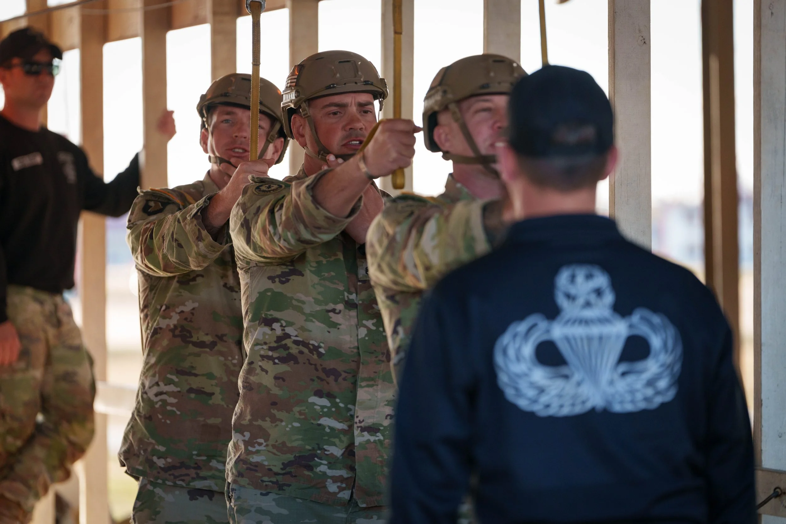 Military personnel participating in a training exercise, with three soldiers wearing camouflage uniforms and helmets, practicing a task under the supervision of a person in a black jacket with a parachute emblem on the back.