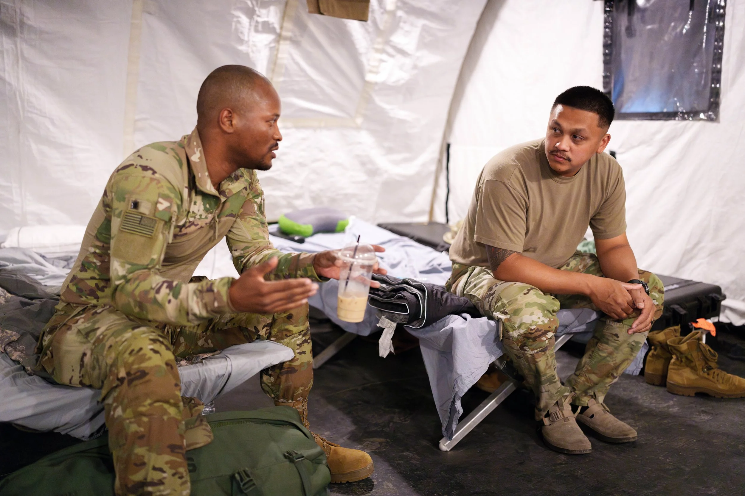 Two soldiers in camouflage uniforms sitting on beds inside a military tent, having a conversation. One is holding a drink and gesturing with his hand, the other is listening attentively.