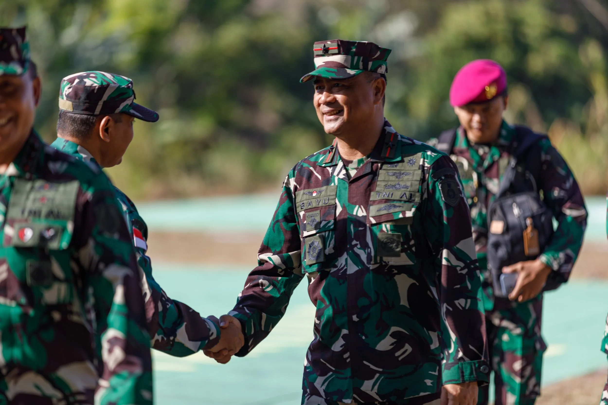 Military officer in camouflage uniform shaking hands with another soldier outdoors, with two other soldiers nearby.