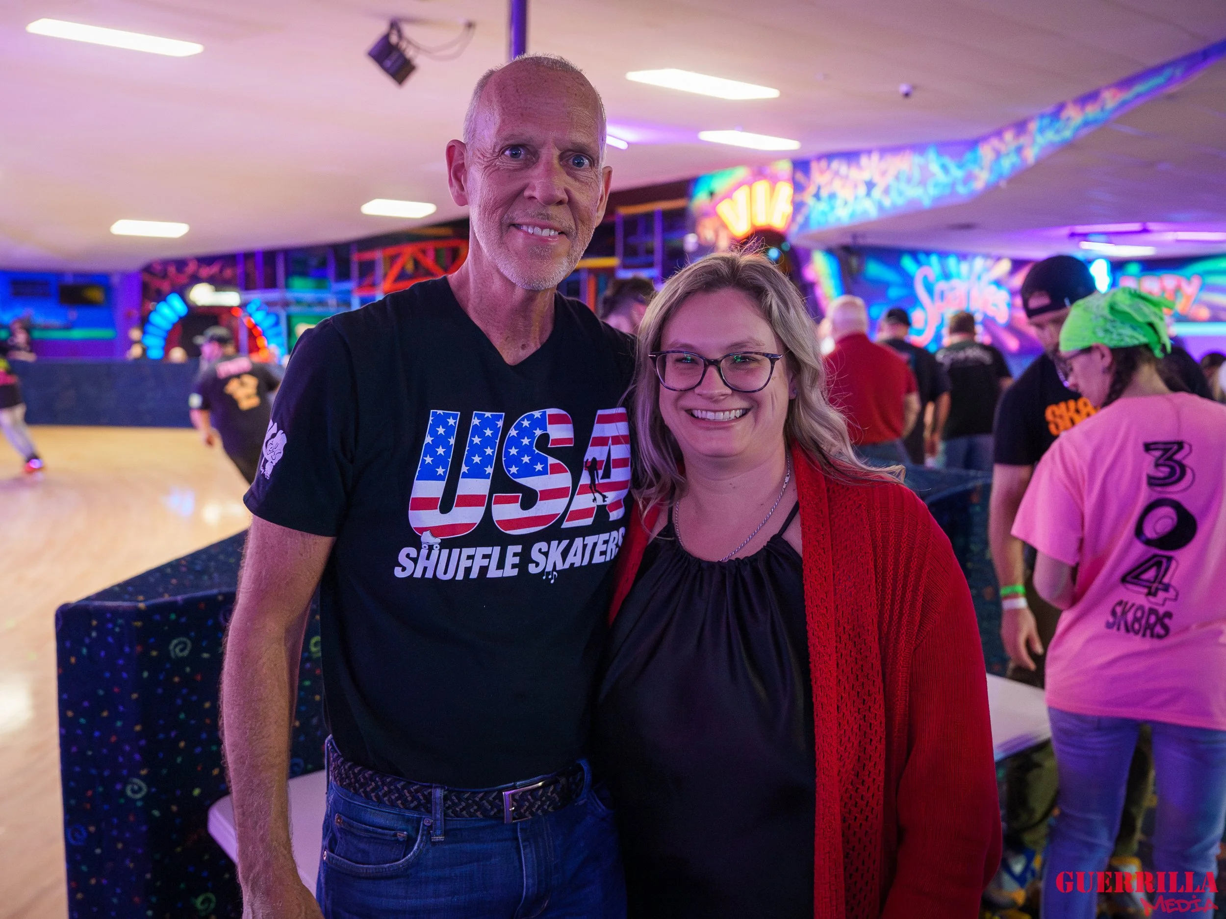 A man and woman smiling at the roller skating rink. The man is wearing a black t-shirt with 'USA Shuffle Skaters' written on it, and the woman is wearing glasses, a black top, and a red cardigan. The background features neon lights and other skaters.