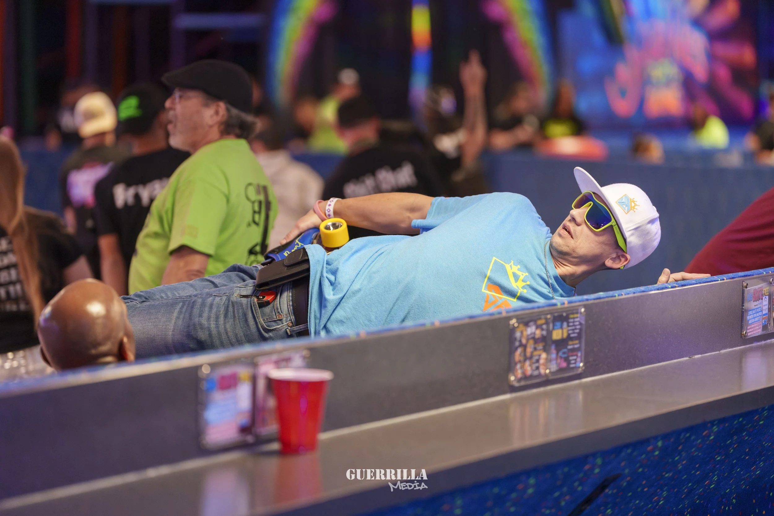 A man wearing sunglasses, a white cap, a light blue t-shirt, and jeans is reclining on a counter at a lively event. He is holding a yellow object and appears to be relaxing, with a crowd and colorful background behind him.