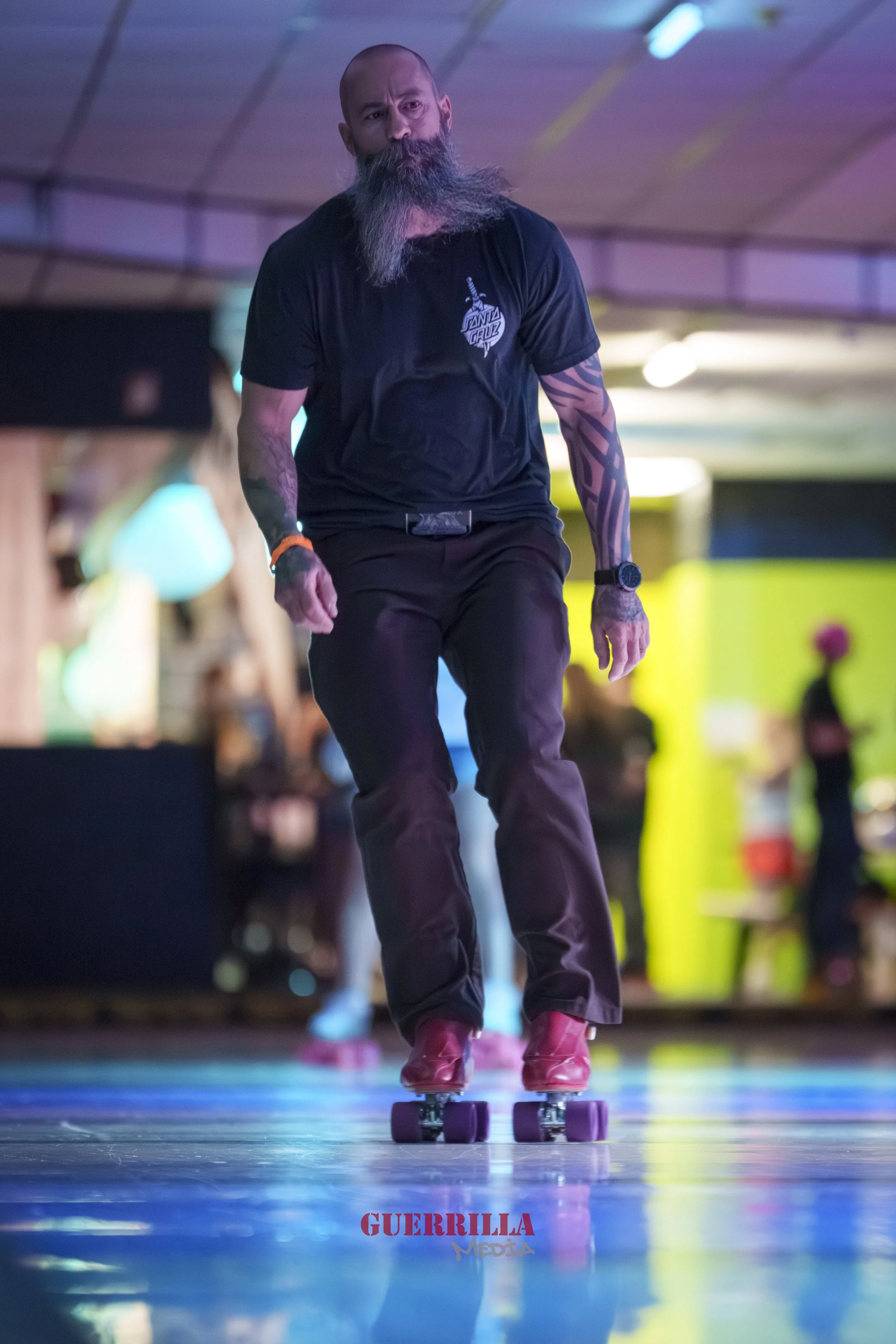 A man with a long beard and a shaved head roller-skates indoors, wearing a black t-shirt and pants, with tattoos on his arms, and a watch on his wrist.