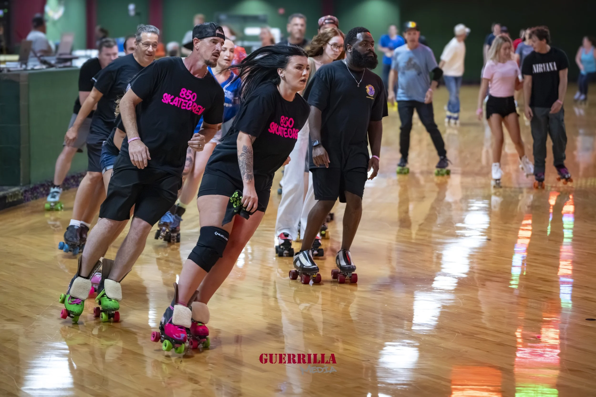 People roller skating in a roller rink, some wearing black T-shirts, with a green walls background and a reflective wooden floor.