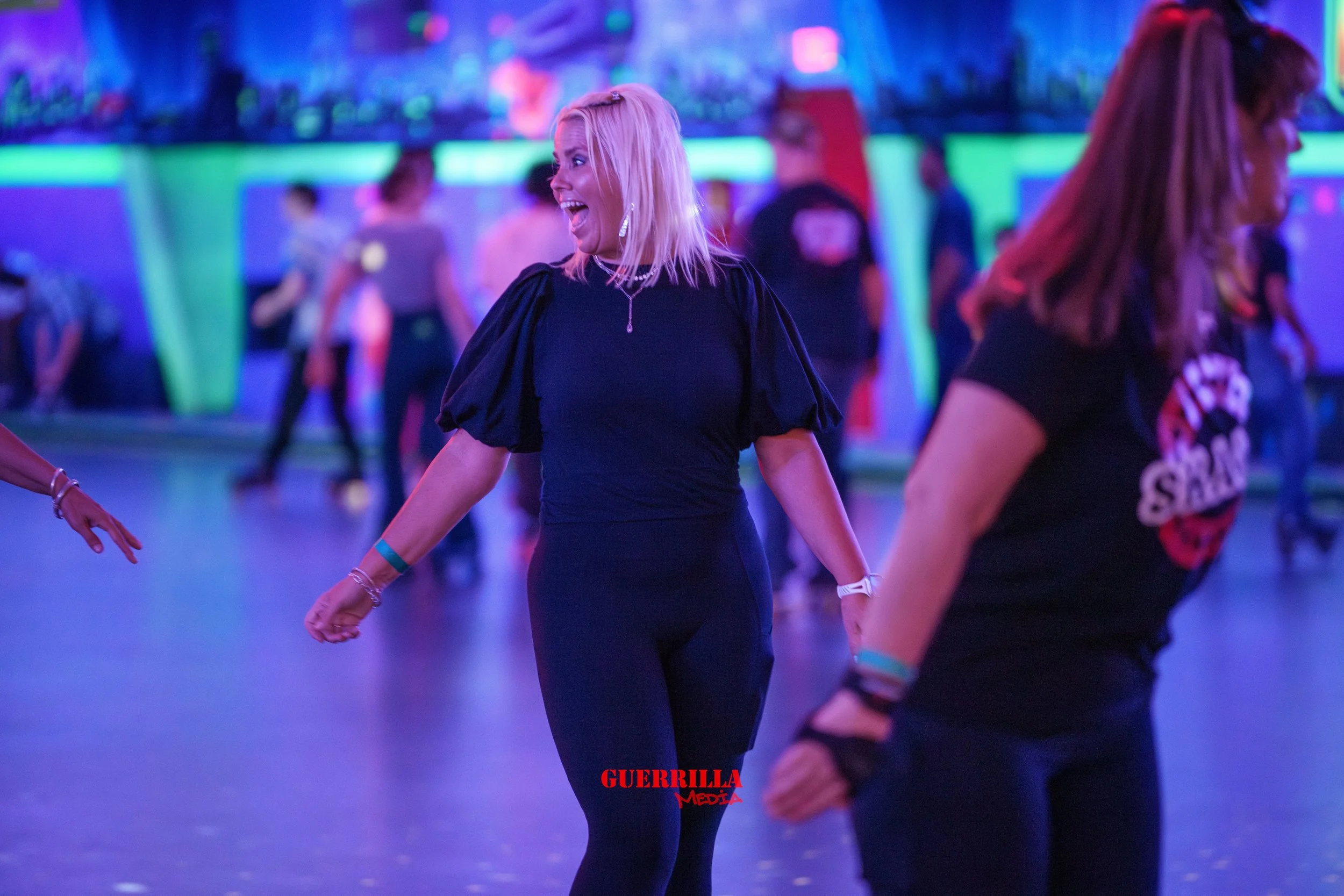 Woman with blonde hair roller skating at an indoor roller rink, smiling and holding hands with others. Neon colorful lighting in background.
