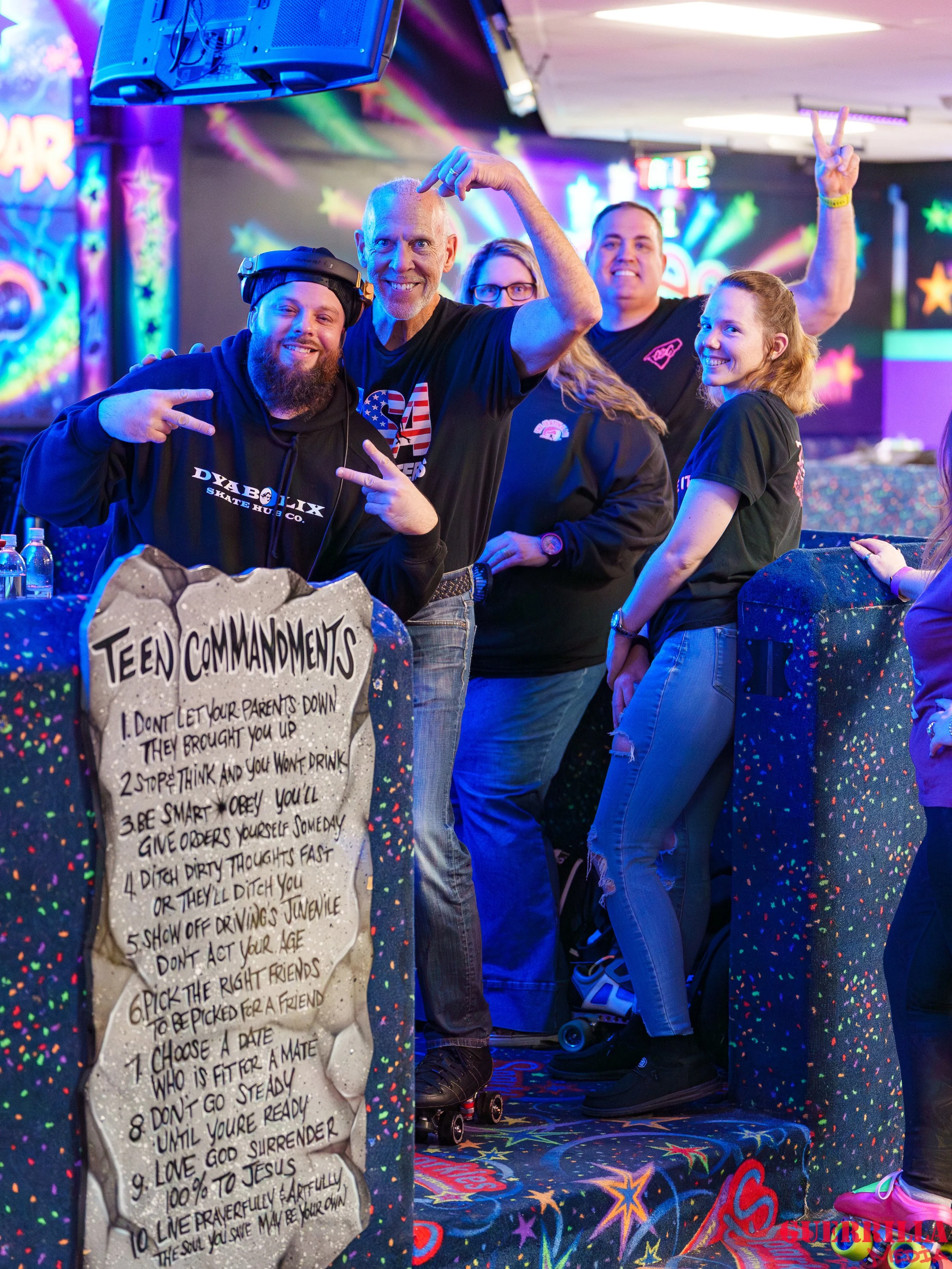 Group of people at a roller skating rink, posing for a photo with colorful neon lights and a Teen Commandments sign in the foreground.