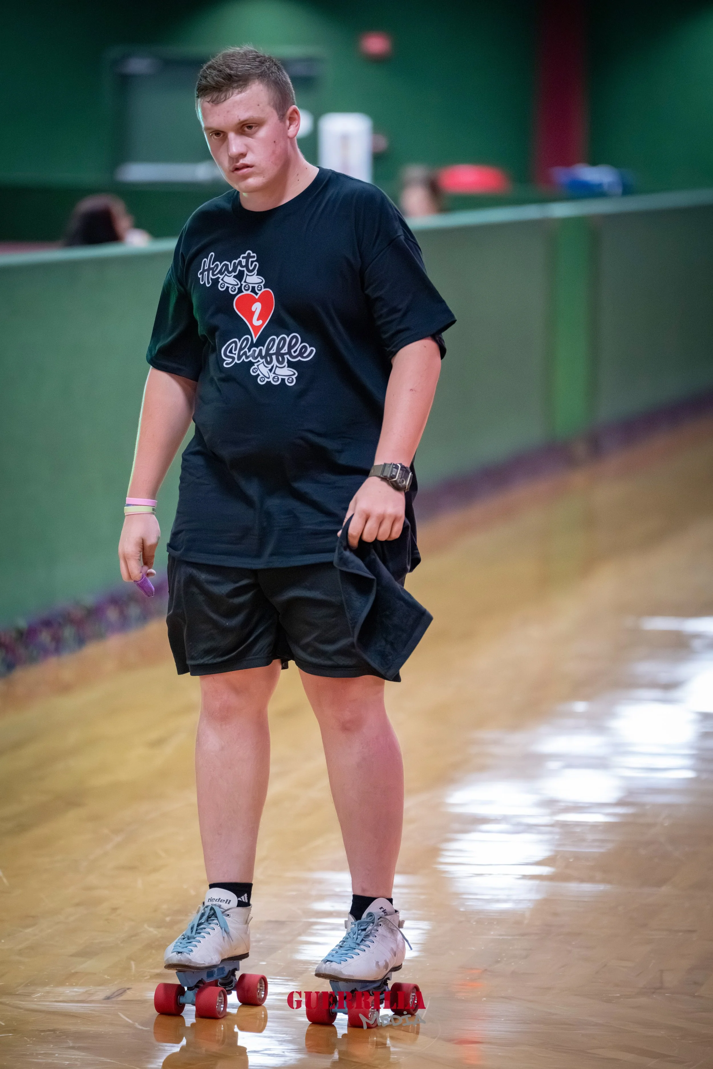A young man roller skating indoors, wearing a black t-shirt with a Heart Shuffle design, black shorts, white roller skates with pink wheels, and holding a jacket in his right hand.