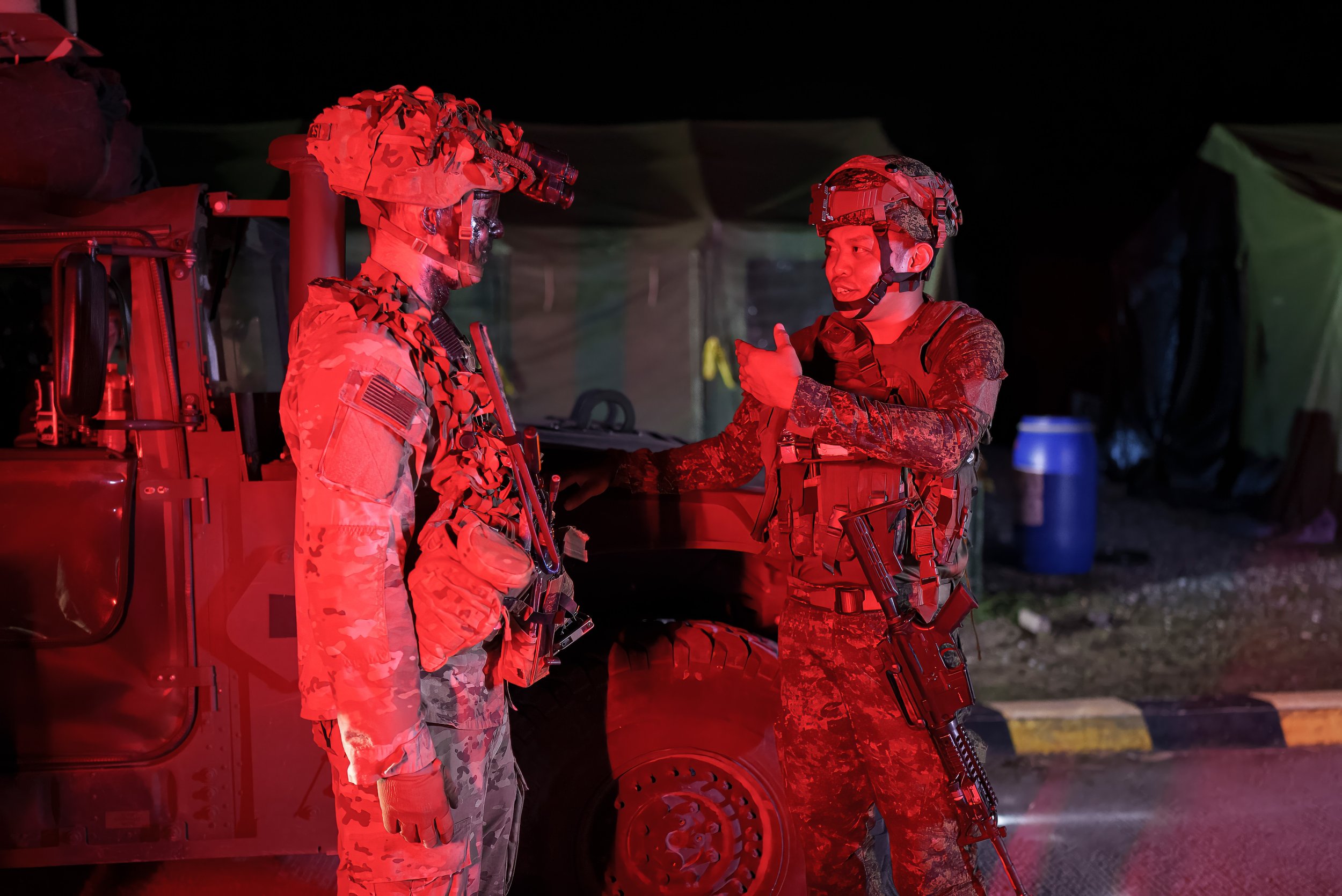 Two soldiers in camouflage uniforms and tactical gear having a conversation at night near a military vehicle, illuminated by red light.