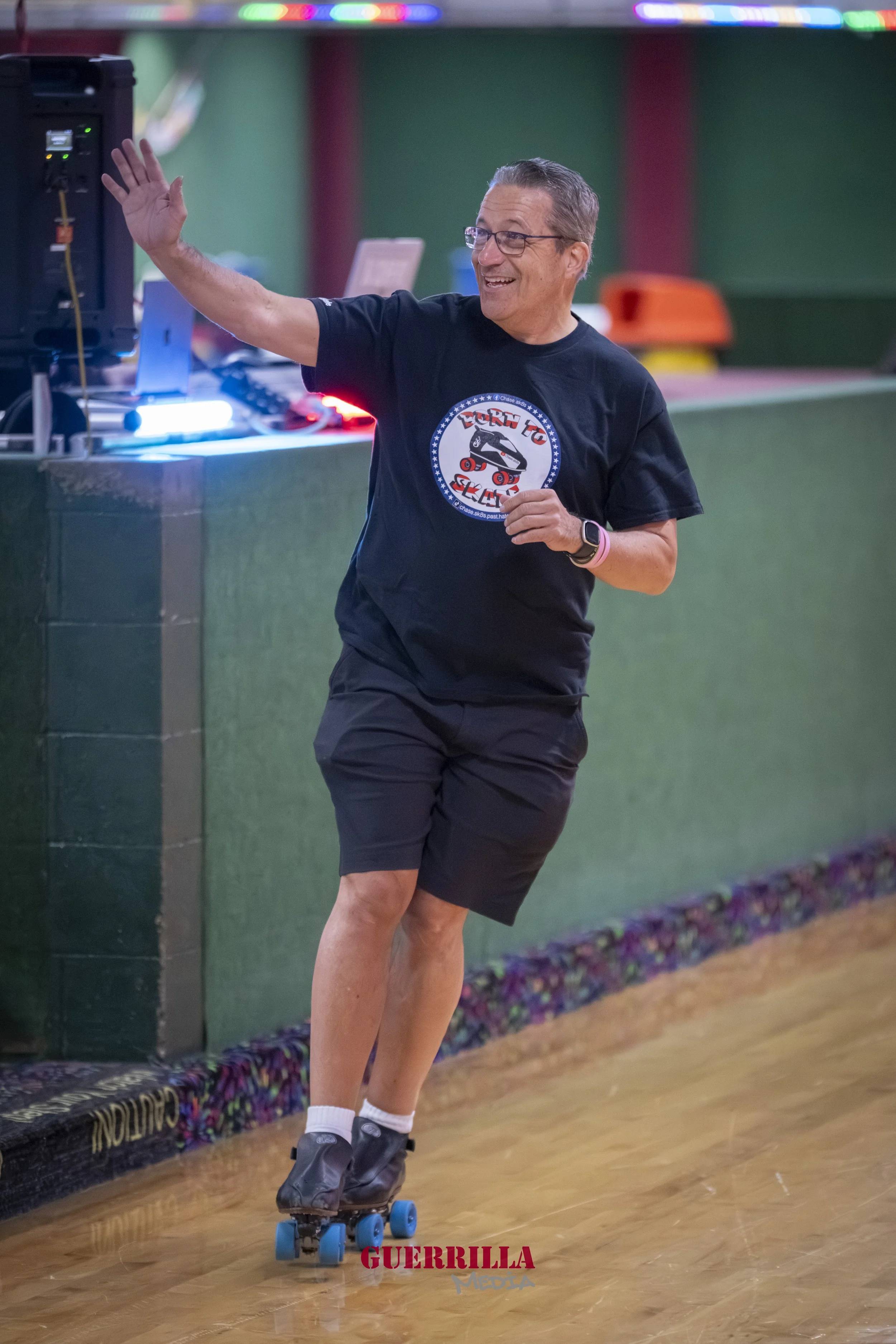 A man roller skating indoors, smiling and waving, wearing a black t-shirt with a roller skate graphic, black shorts, and white socks.