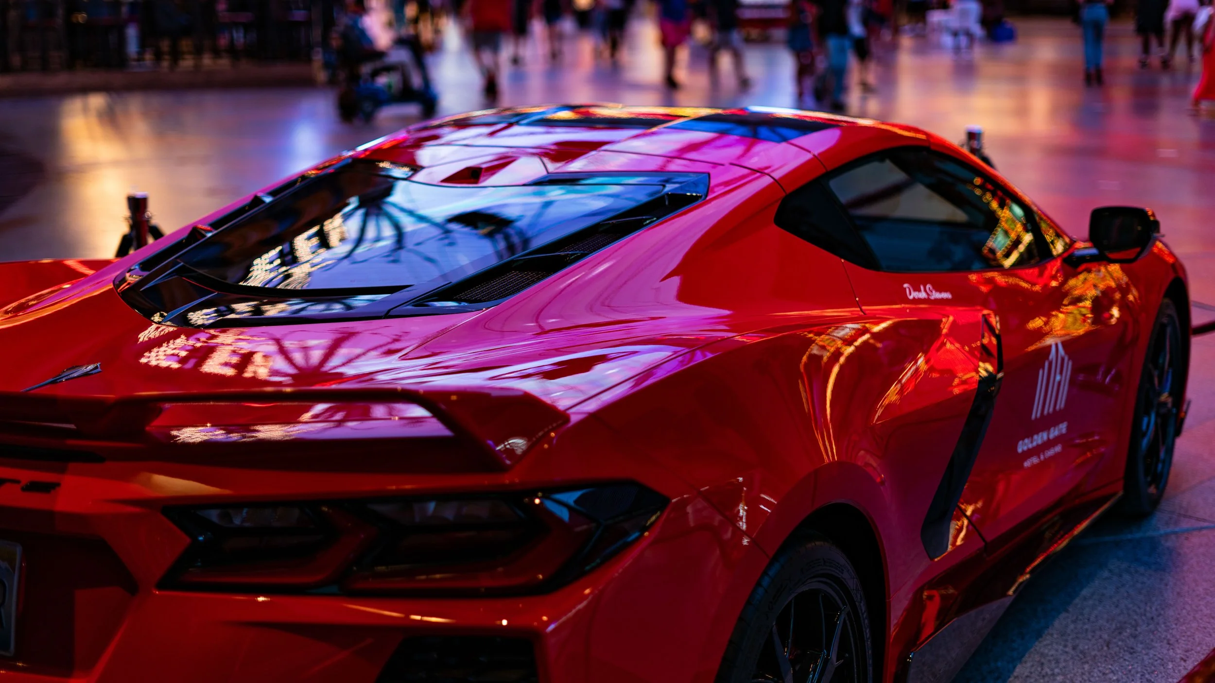 A shiny red sports car parked in an indoor area with a blurred background of people and lights.