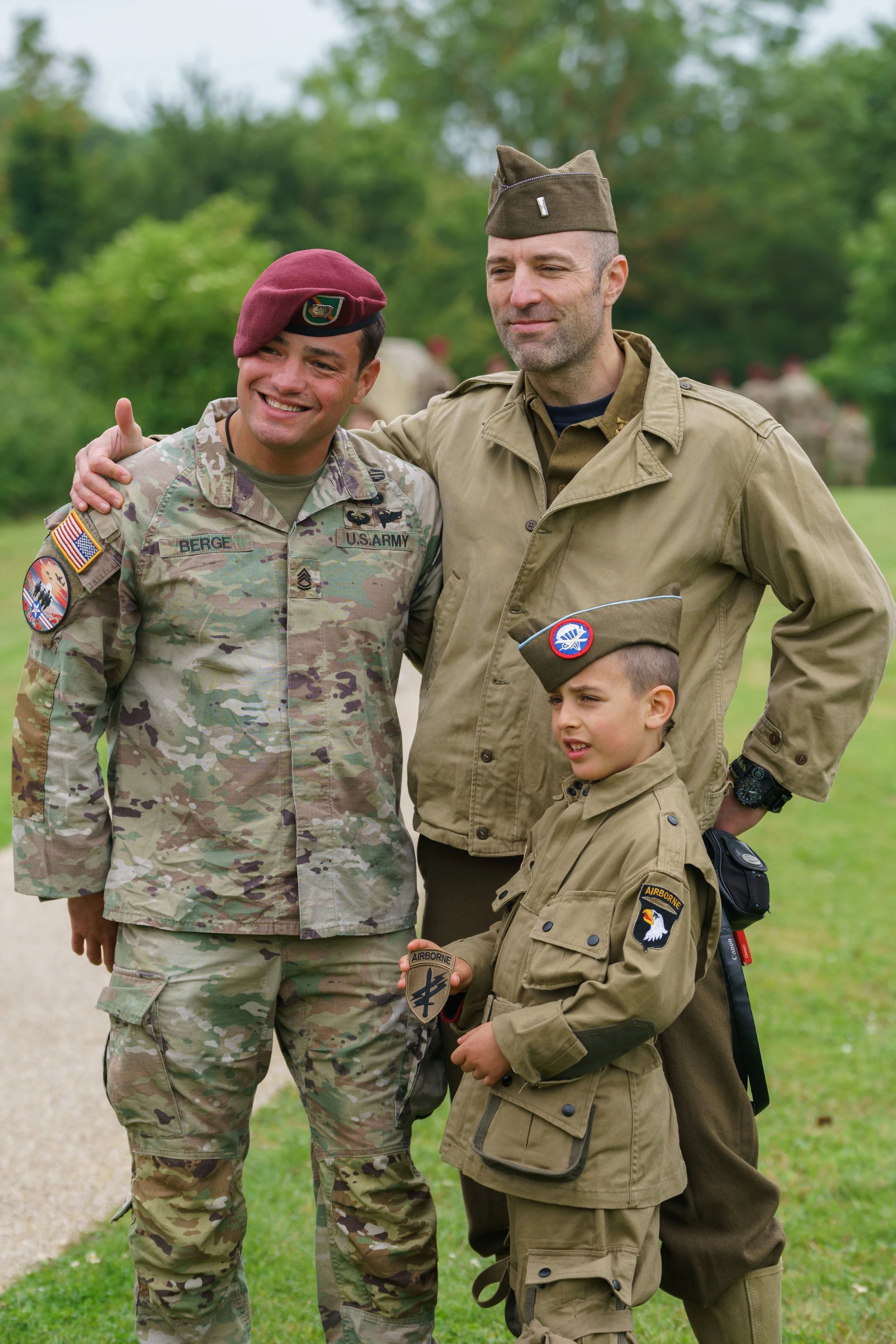 Three people in military uniforms smiling outdoors, with one young boy holding a military badge, in a grassy park with trees.