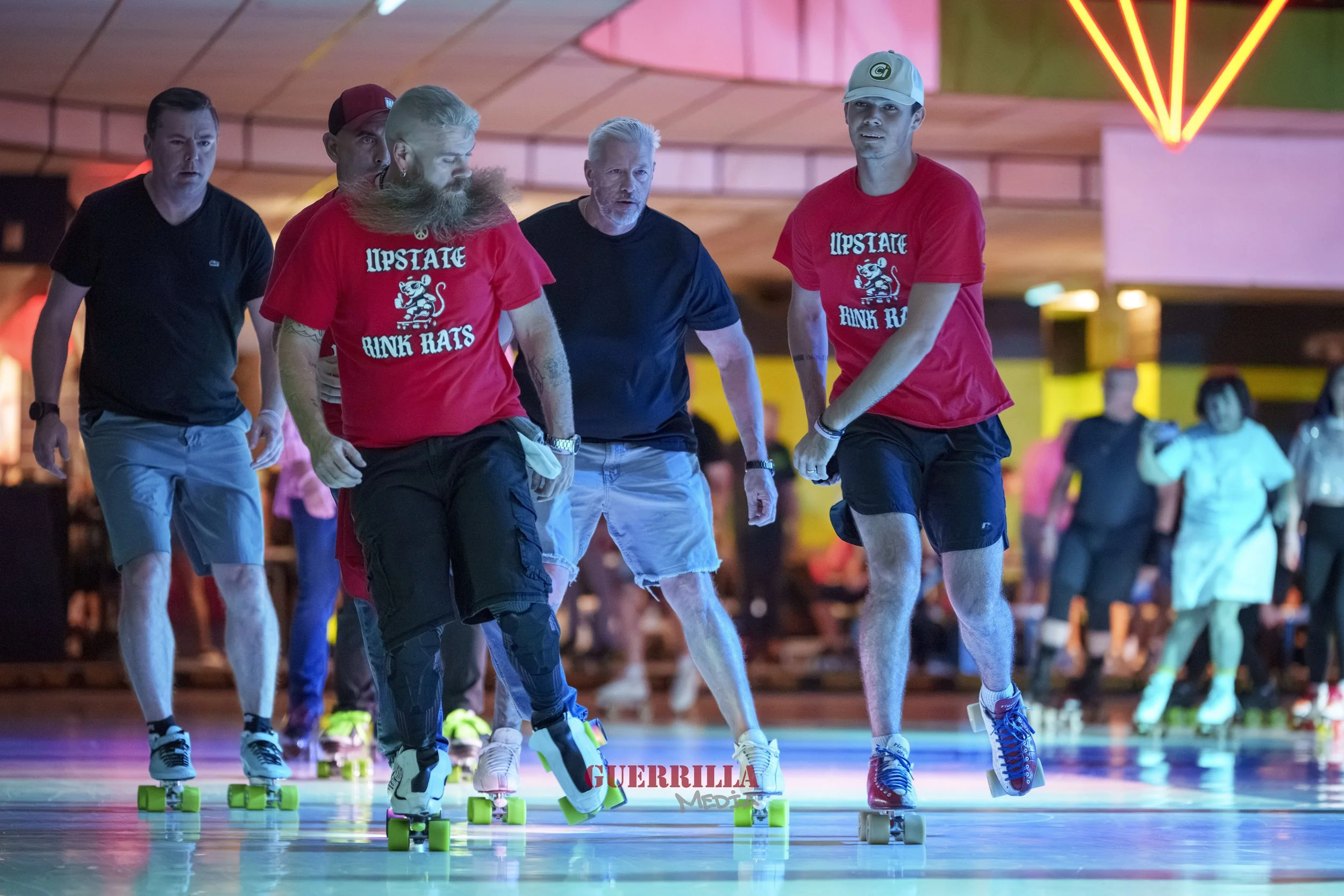 People roller skating at an indoor rink with colorful lighting and a wooden ceiling, some wearing red shirts with 'Upstate Rink Rats' logos.