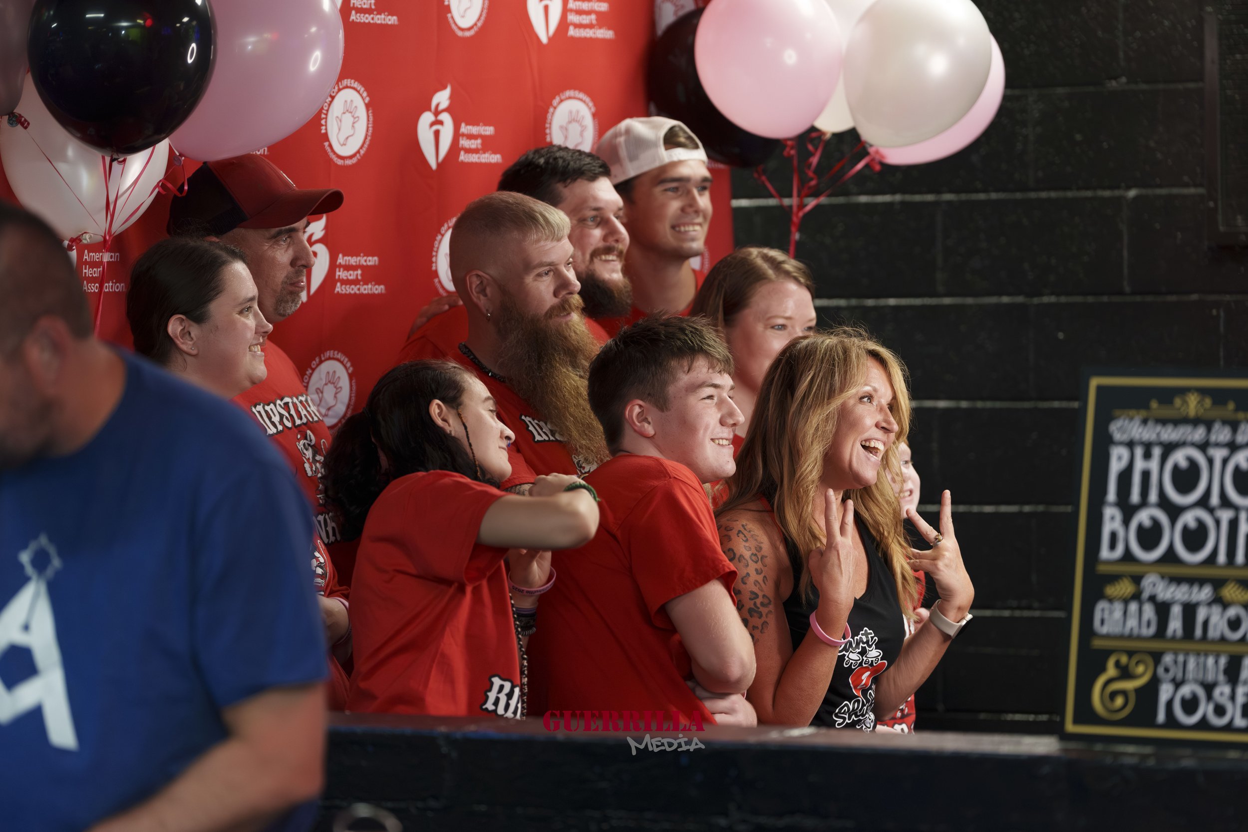 Group of people at an American Heart Association event, posing for a photo with balloons and a red backdrop, some smiling and making peace signs.
