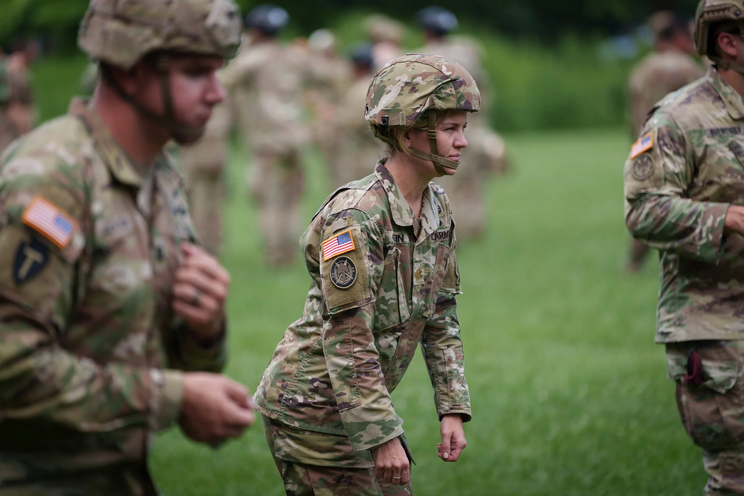 Group of soldiers in camouflage uniforms standing outdoors on a grassy field, with one female soldier in the center looking serious, wearing a helmet and military patches.