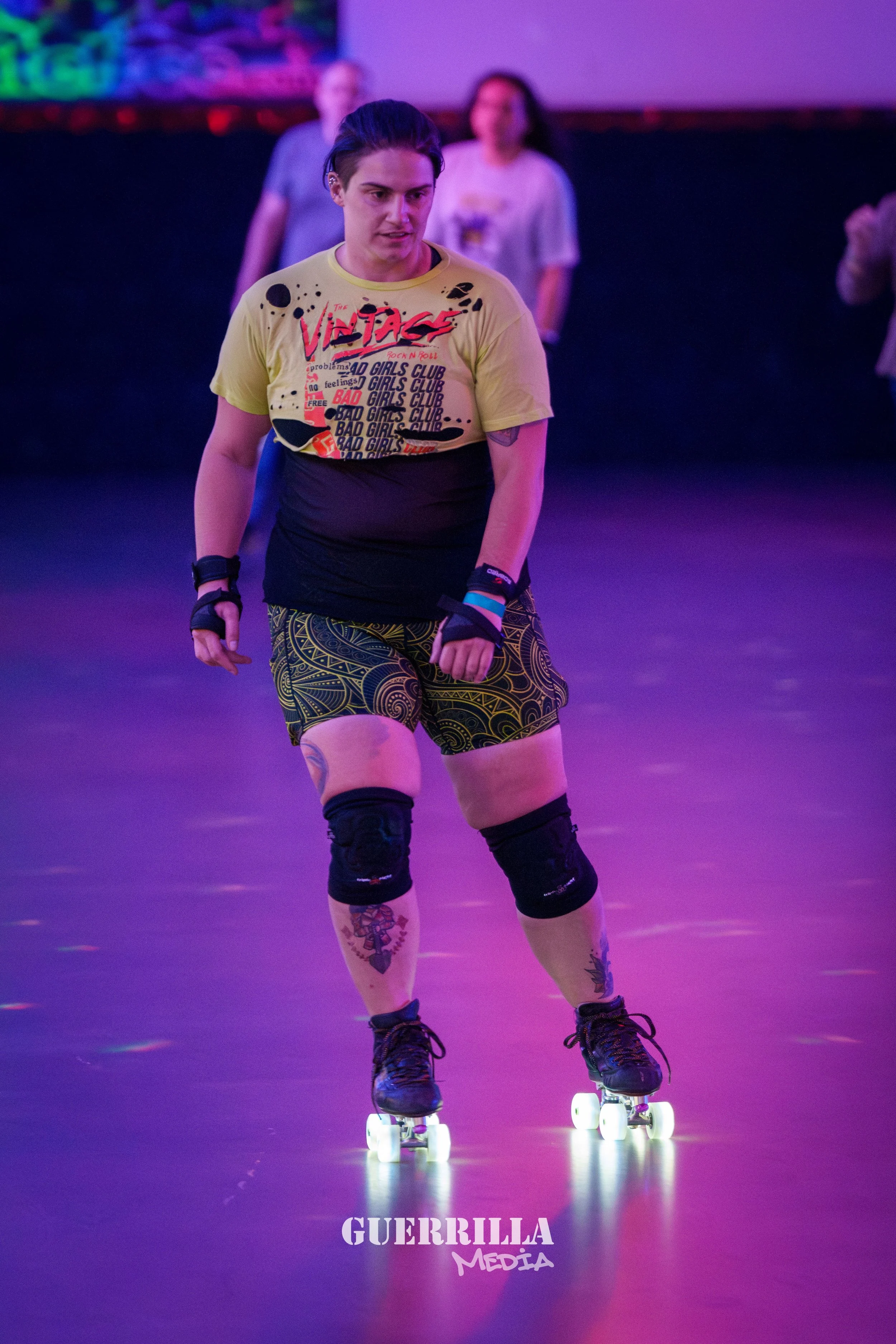 A person roller skating indoors under purple lighting, wearing a yellow and black shirt, patterned shorts, knee and wrist pads, and tattoos on their legs.