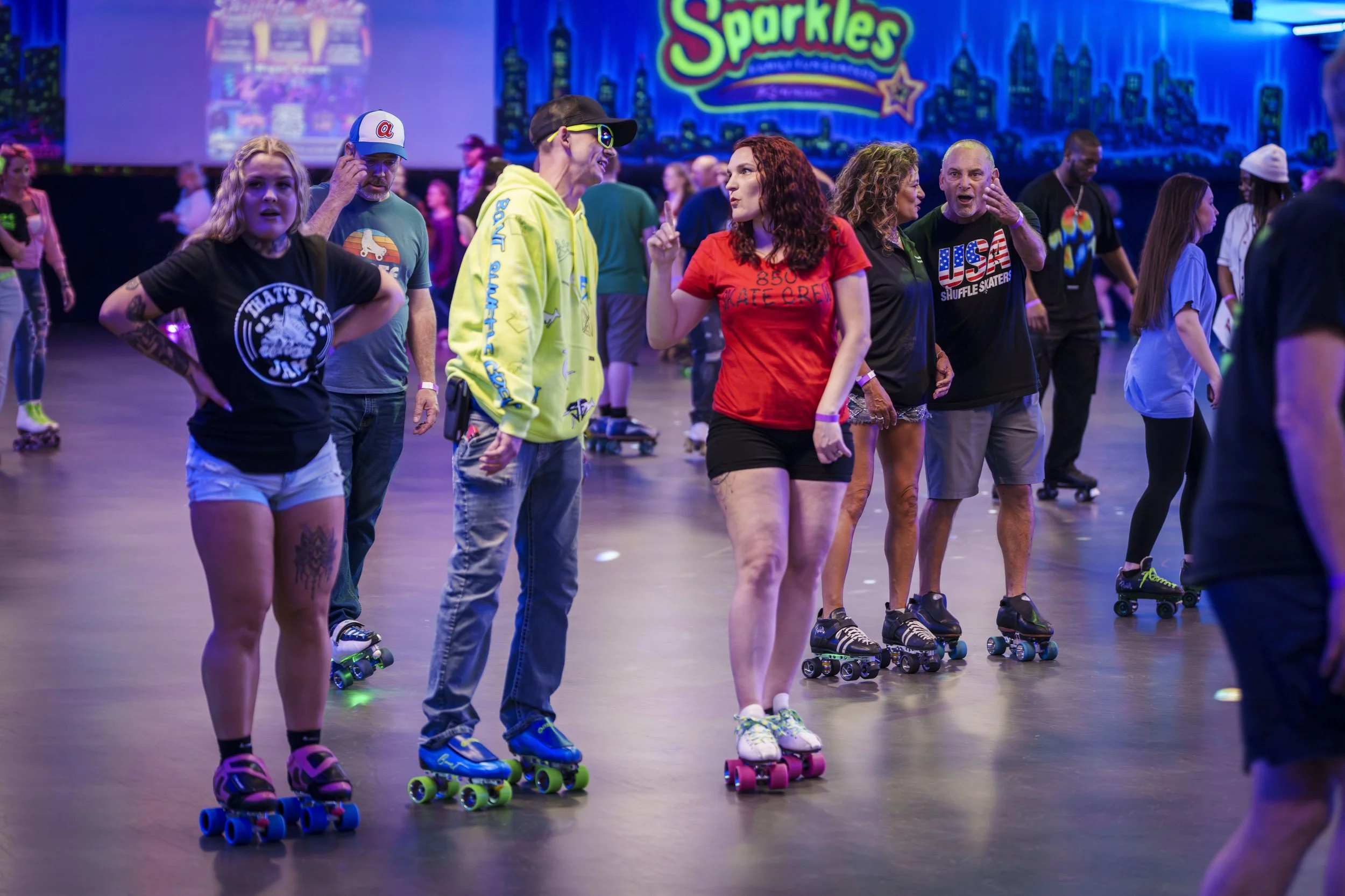 People roller skating in an indoor rink with colorful neon lighting and a cityscape backdrop displaying the word 'Sparkles'.