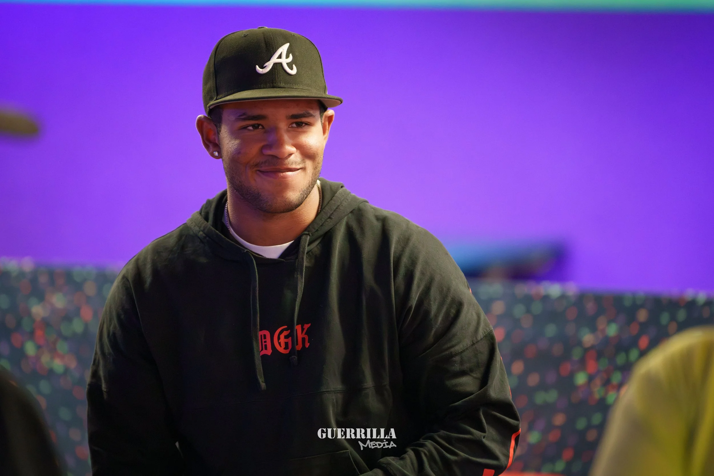 Young man wearing a black cap with a white 'A' logo, black hoodie, earrings, and a small chain, smiling in front of a colorful, blurred background.