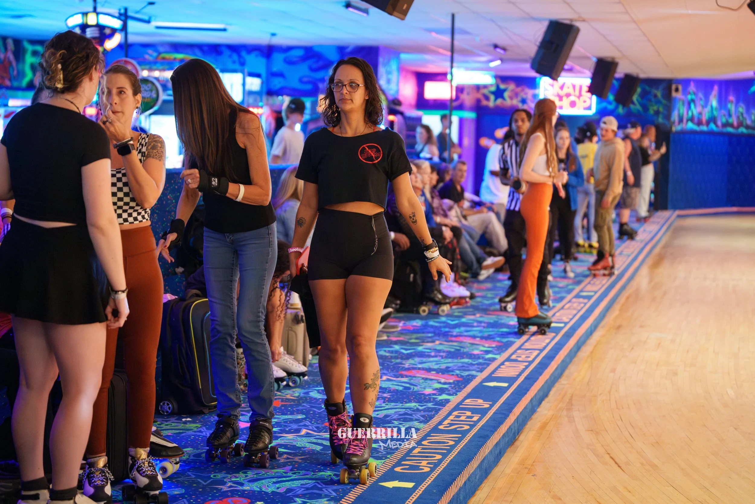 People roller skating inside a roller rink with colorful neon lights, seated spectators, and playful surf-themed wall murals.