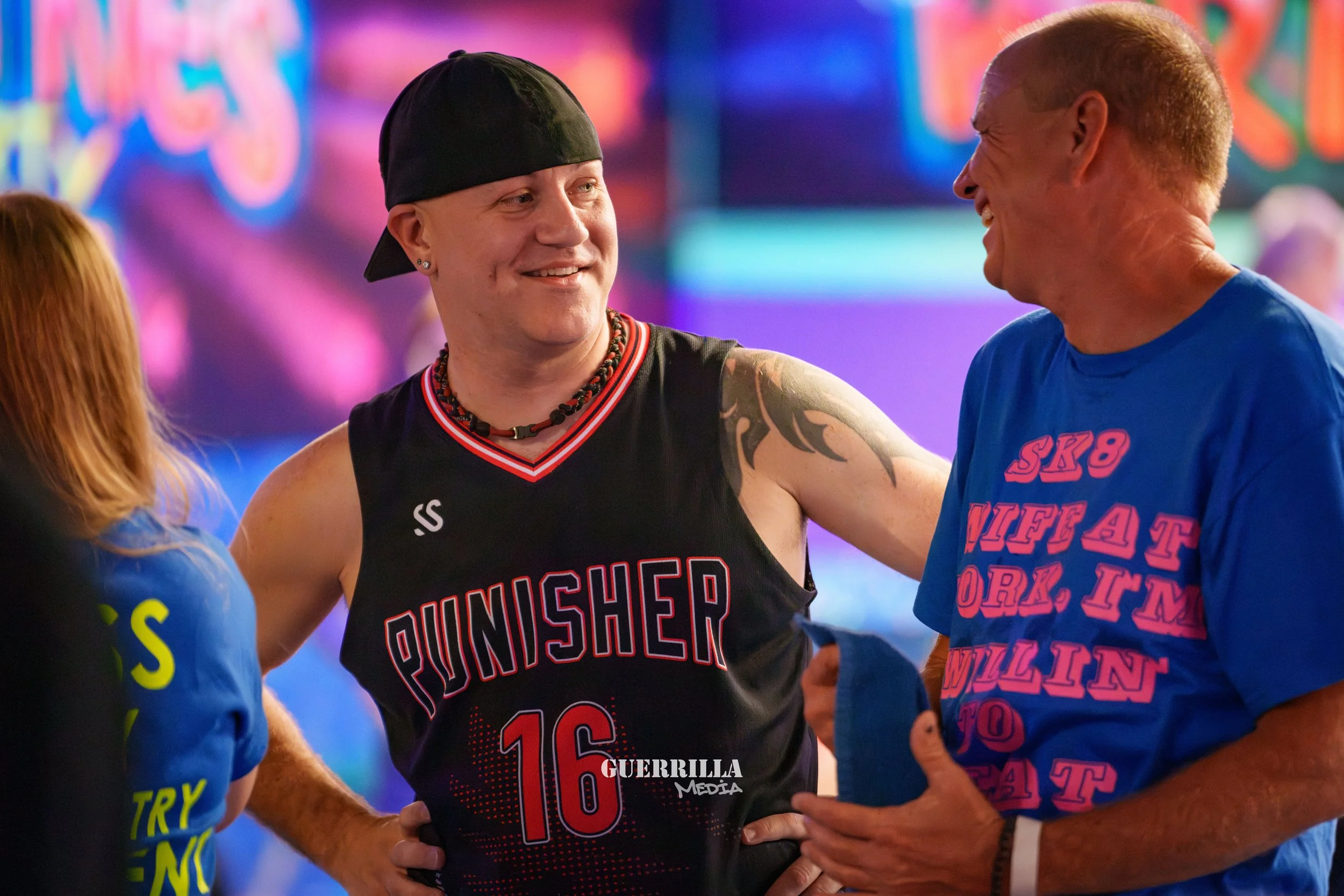 A woman in a black sports jersey with 'Punisher' and the number 16, smiling and talking with a man in a blue shirt, in a colorful indoor setting.