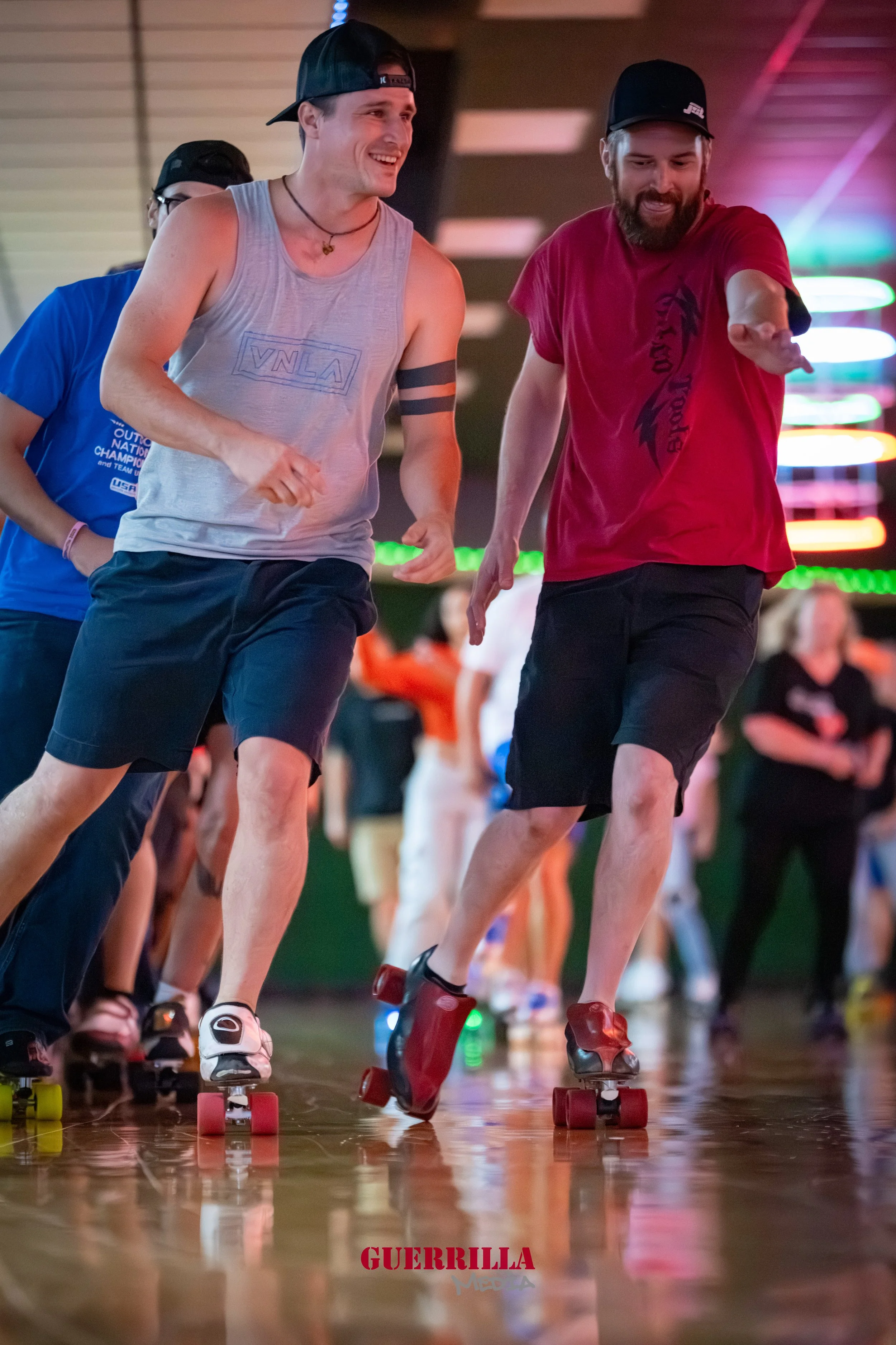 Two young men rollerskating indoors at a skating rink, smiling and having fun, with a crowd in the background and colorful lights.