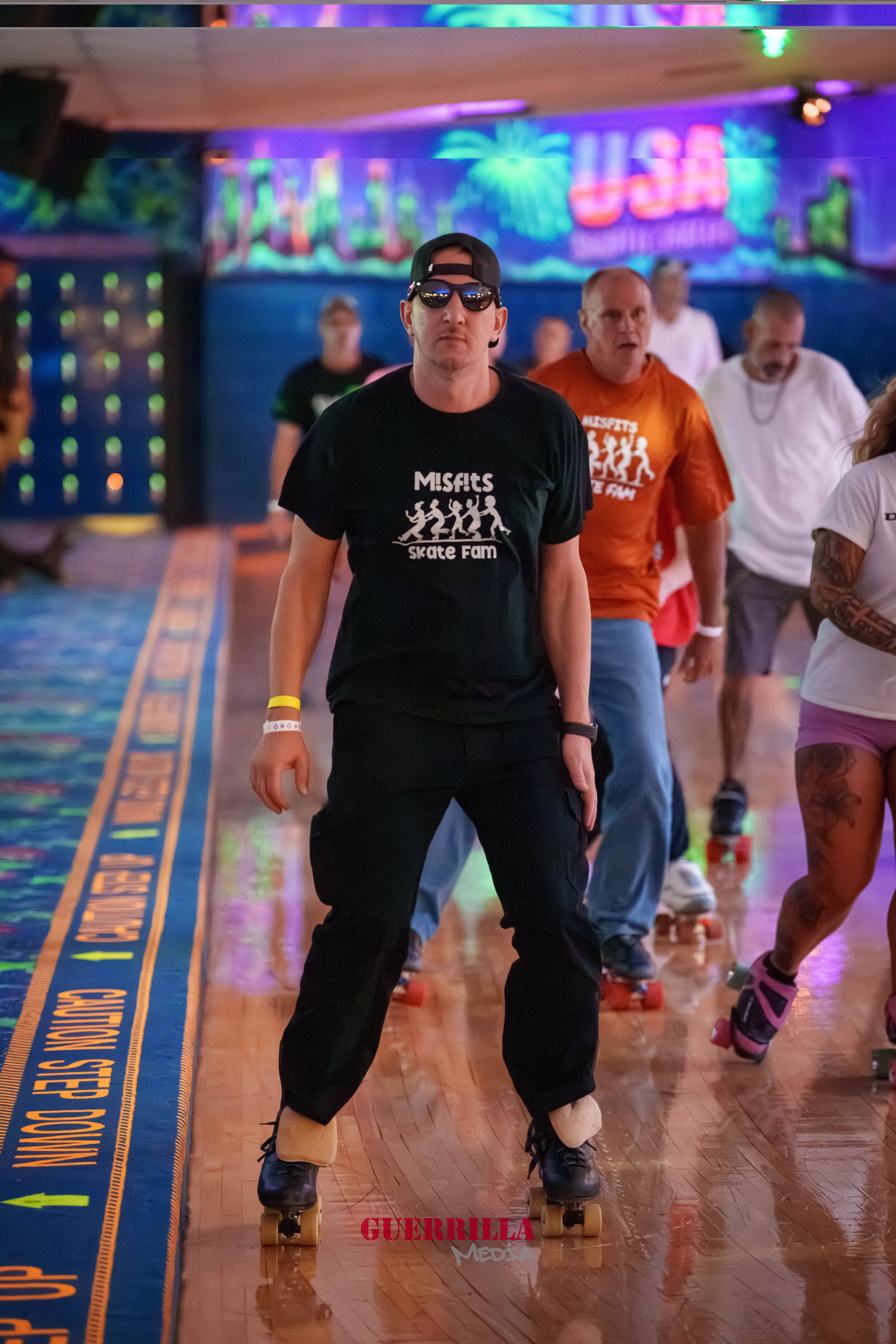 A group of people roller skating indoors, some wearing Misfits band t-shirts, with colorful neon lights and painted wall murals in the background.