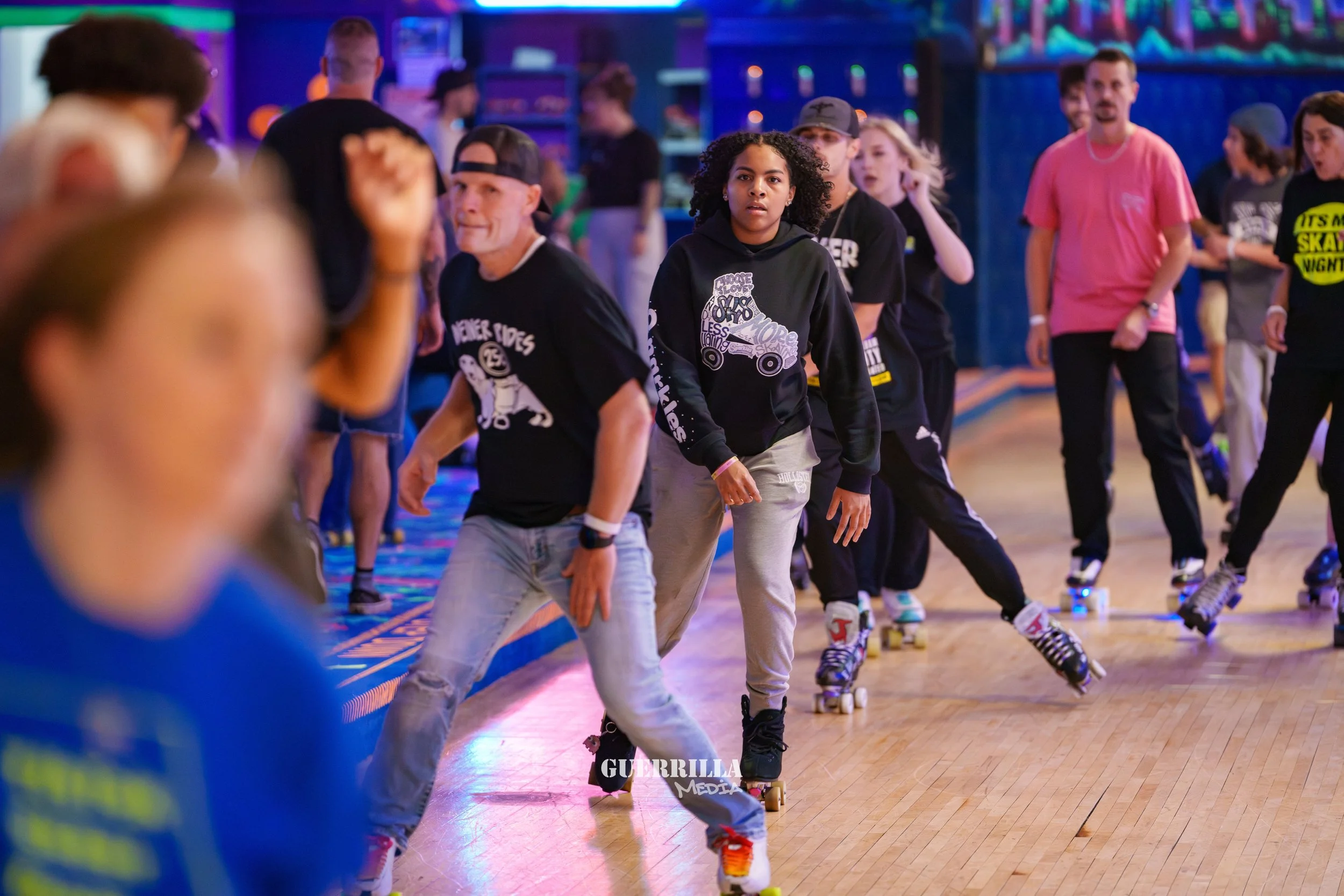 People roller skating in an indoor rink under colorful neon lights, with some individuals looking towards the camera and others skating or watching.