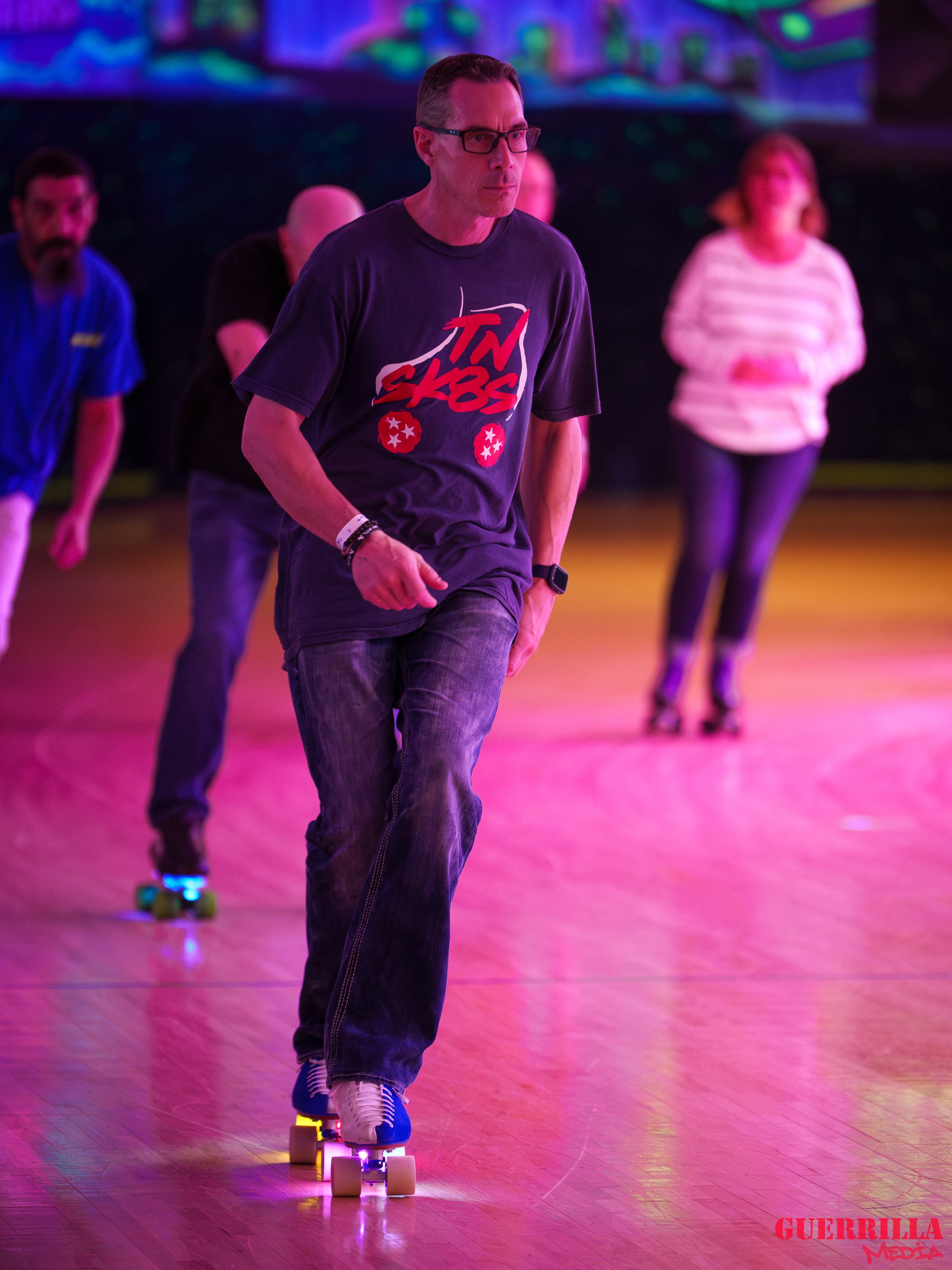 People roller skating indoors under colorful neon lights, with a man in the foreground wearing glasses, a black shirt, and jeans.