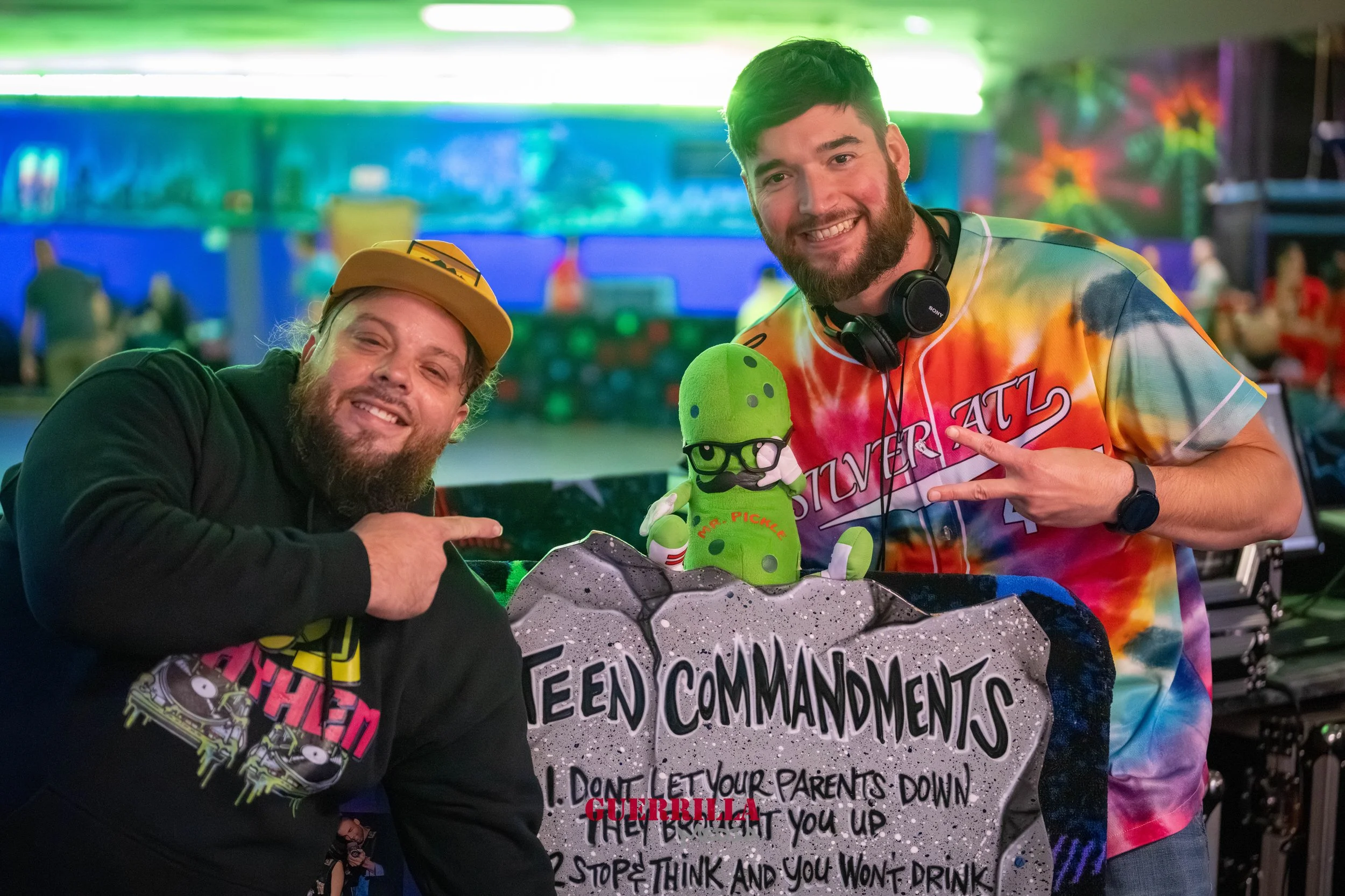 Two men in colorful clothing pointing at a plush toy of 'Mr. Pickle' wearing glasses and a mustache, in front of a sign with 'Teen Commandments'. The background shows a lively, neon-lit indoor space.