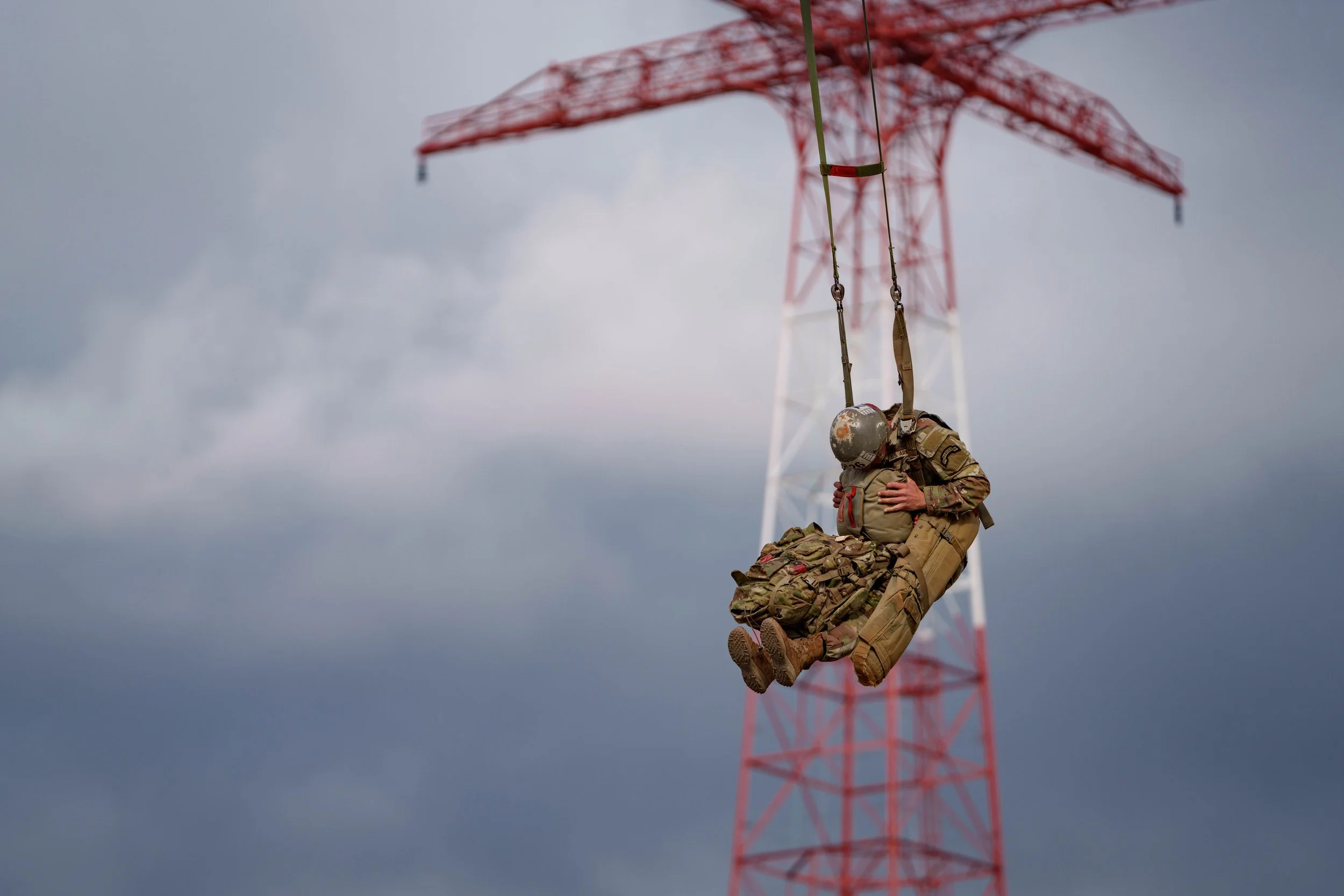 A soldier in military uniform and helmet hanging from a rescue harness in the air near a large red and white communication tower against a cloudy sky.