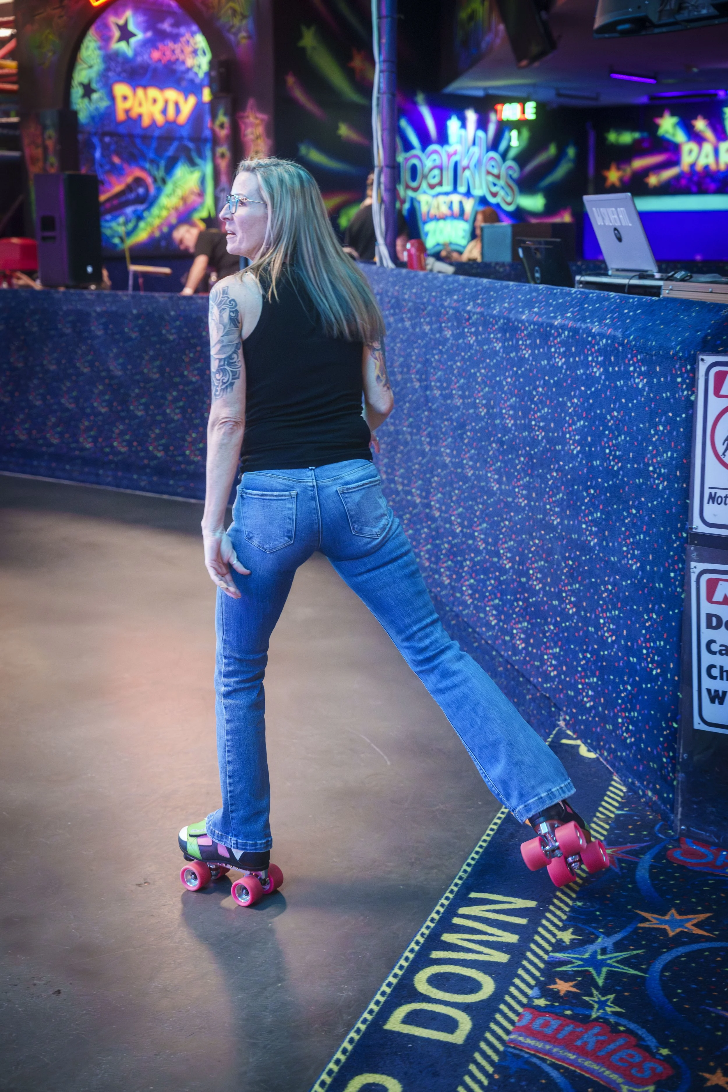 A woman with long blonde hair, tattoos on her arms, wearing glasses, a black sleeveless top, and blue jeans, is roller skating inside a neon-lit roller disco. She is standing on one leg with the other extended behind her, on pink roller skates, next 
