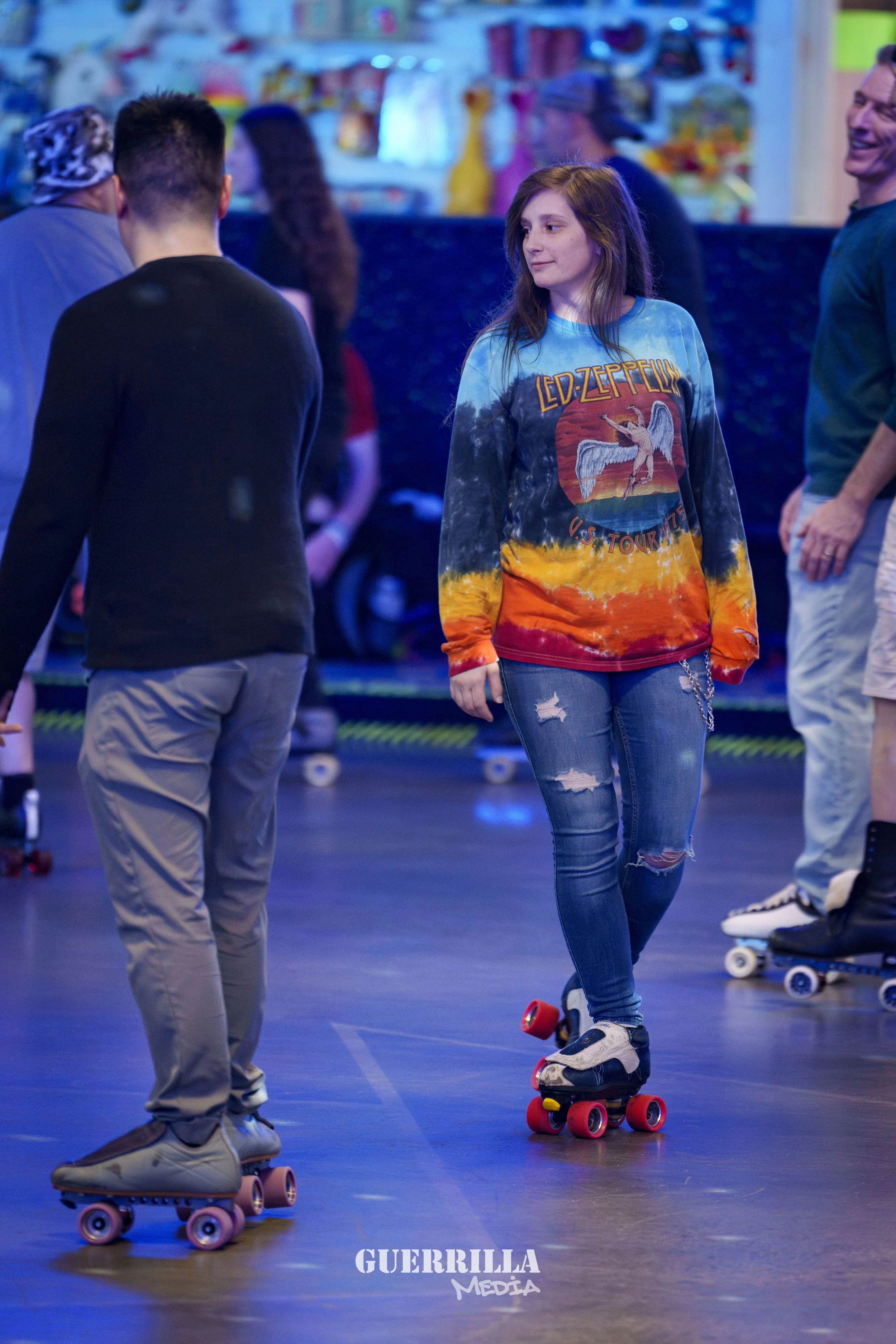 A young woman with long reddish hair roller skating indoors, wearing a Led Zeppelin T-shirt, ripped jeans, and black skate shoes. She is smiling slightly and looking to her right. Other people are in the background, also skating or standing around. T