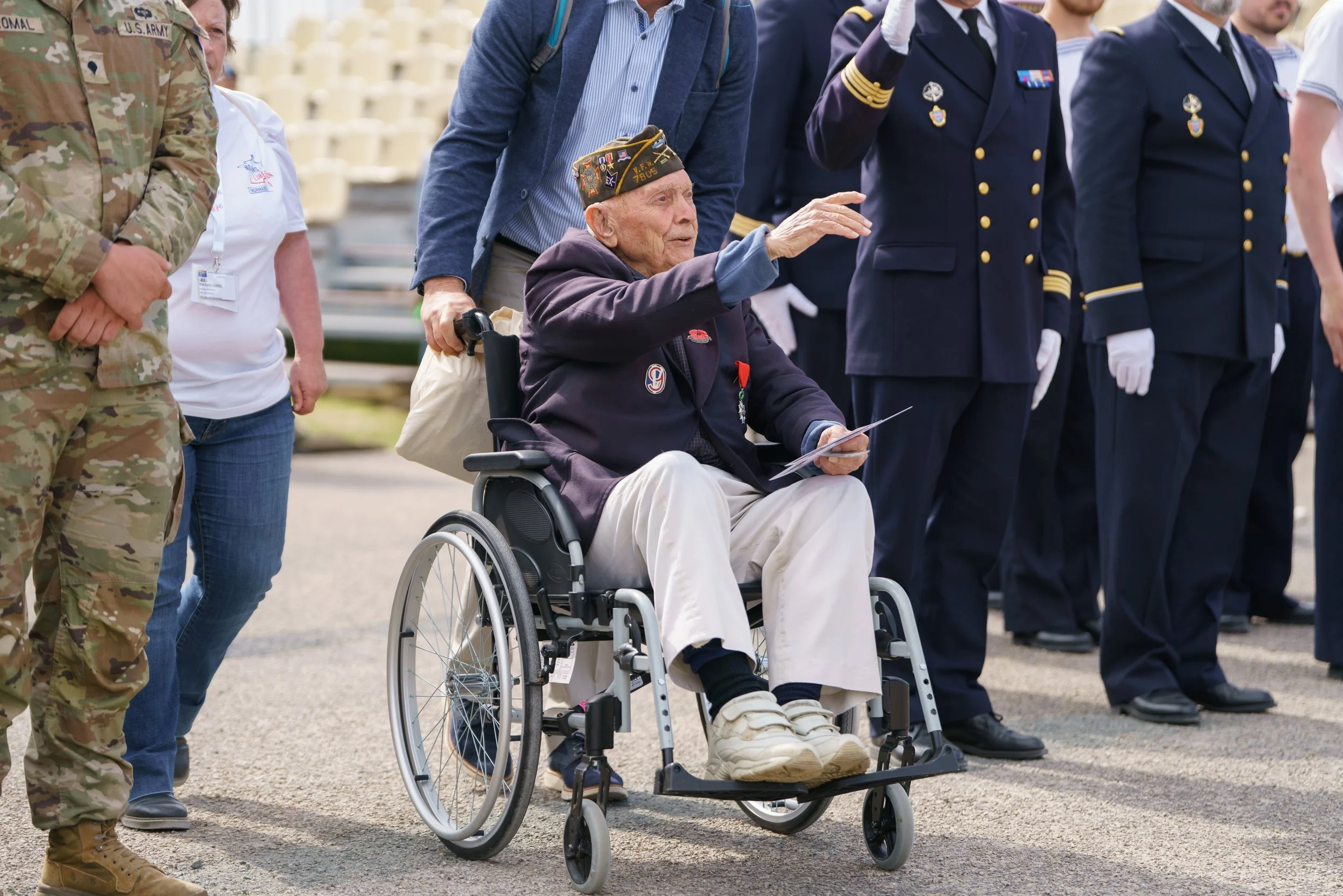 An elderly man in a wheelchair, dressed in a dark blazer and light-colored pants, wears a veteran hat with patches. He is holding a photo in one hand and waving with the other. Several people in military uniform and casual clothing stand around him a