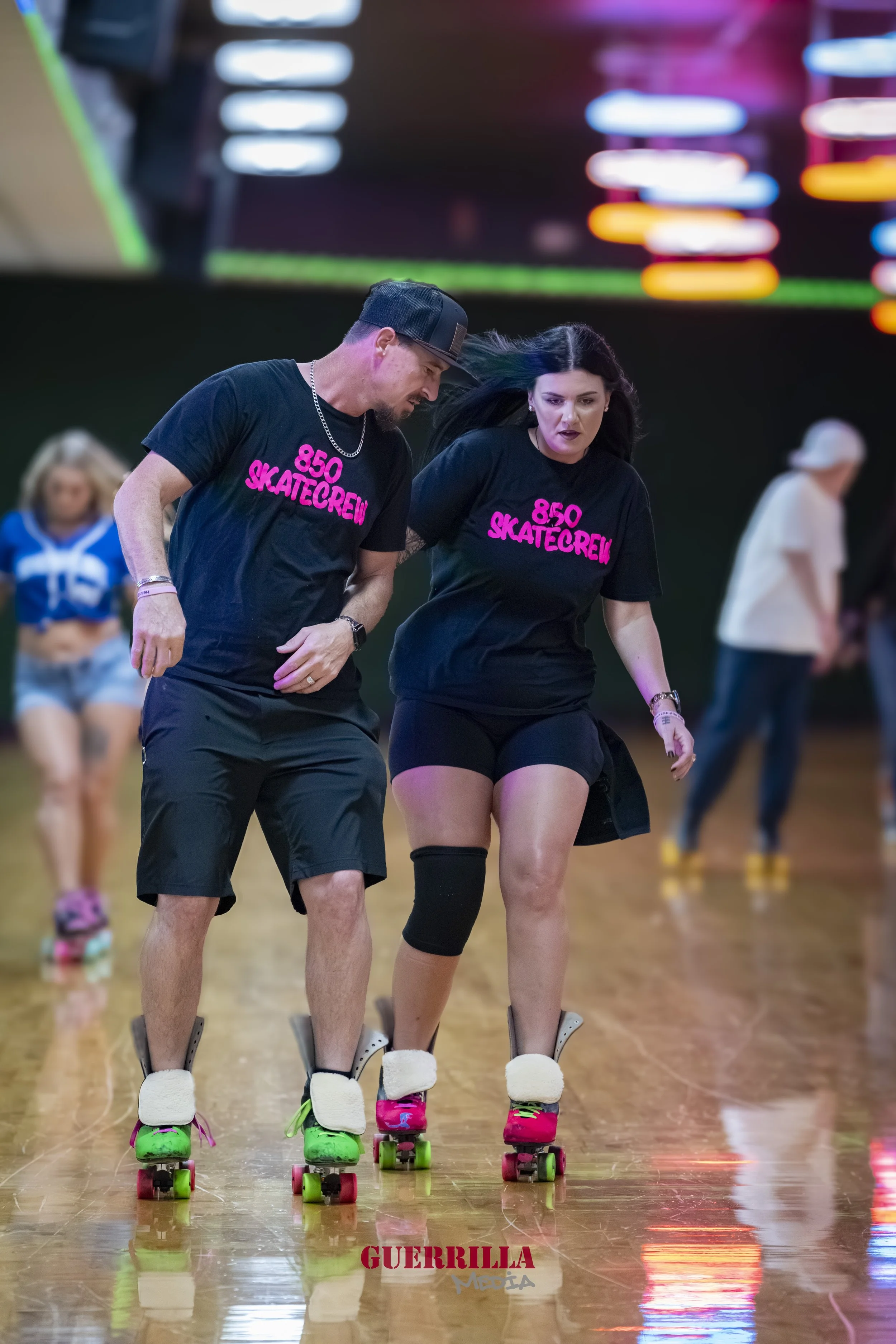 Two people roller skating on a rink, wearing matching black t-shirts with pink text, with colorful lights and other skaters in the background.
