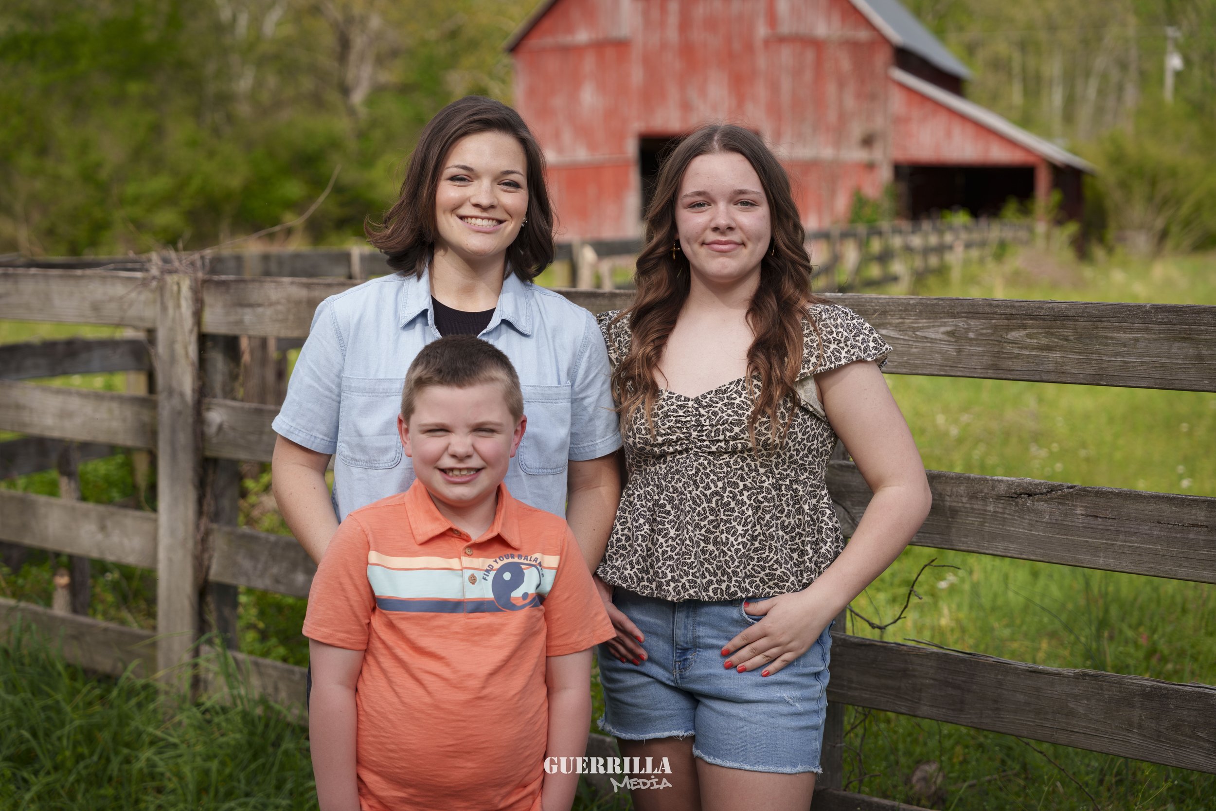 Three children standing outdoors in front of a wooden fence with a red barn in the background. The children are smiling and posing for the photo.