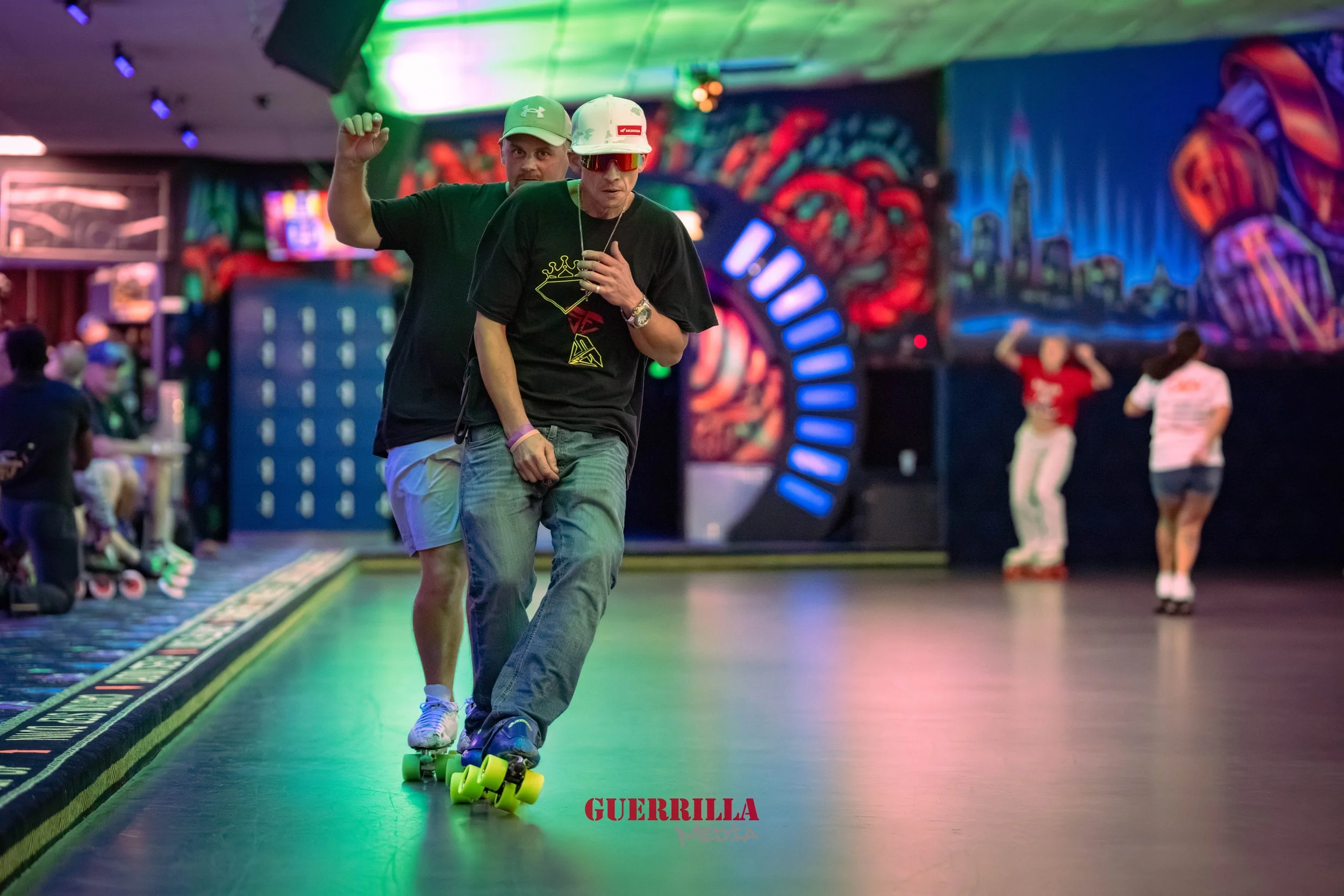 Two men roller skating inside an indoor skating rink with neon lights and colorful graffiti on the walls. The man in the front is wearing a black t-shirt, blue jeans, and sunglasses, while the man behind him is wearing a black t-shirt and light-color