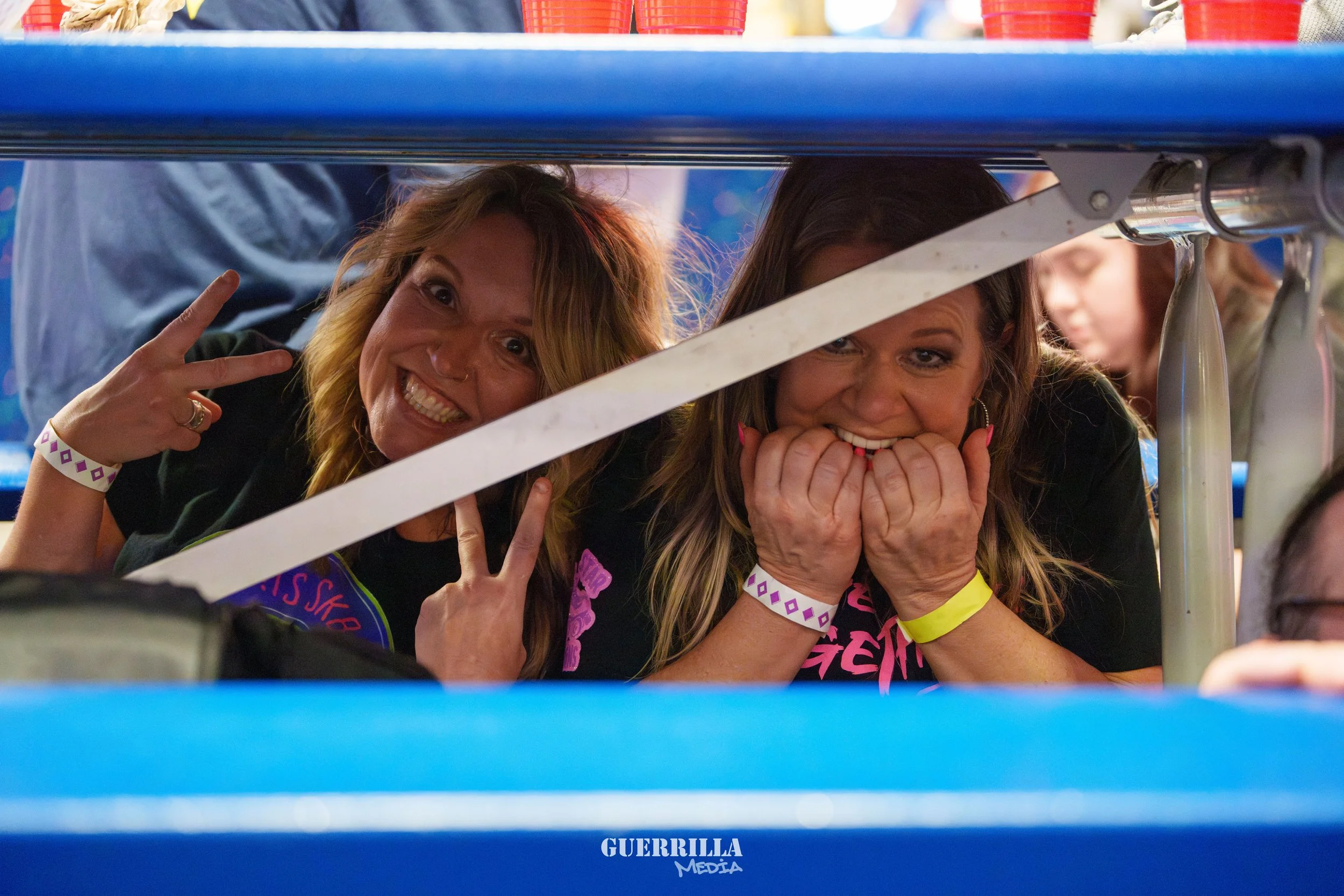 Two women smiling and making playful gestures at an event, seen from below through a blue barrier.