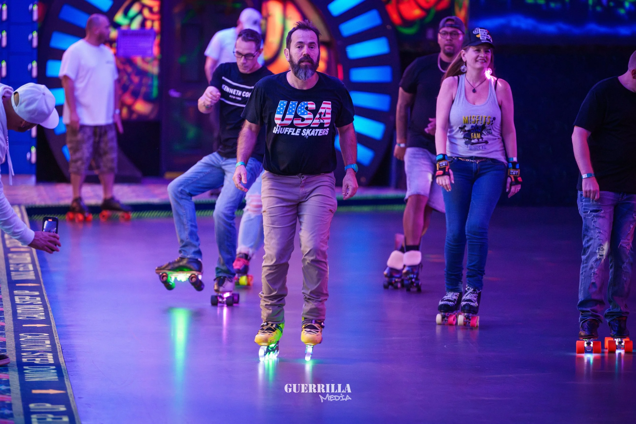 A group of people roller skating indoors under colorful neon lights, some wearing casual clothes and wristbands, with a digital display in the background.