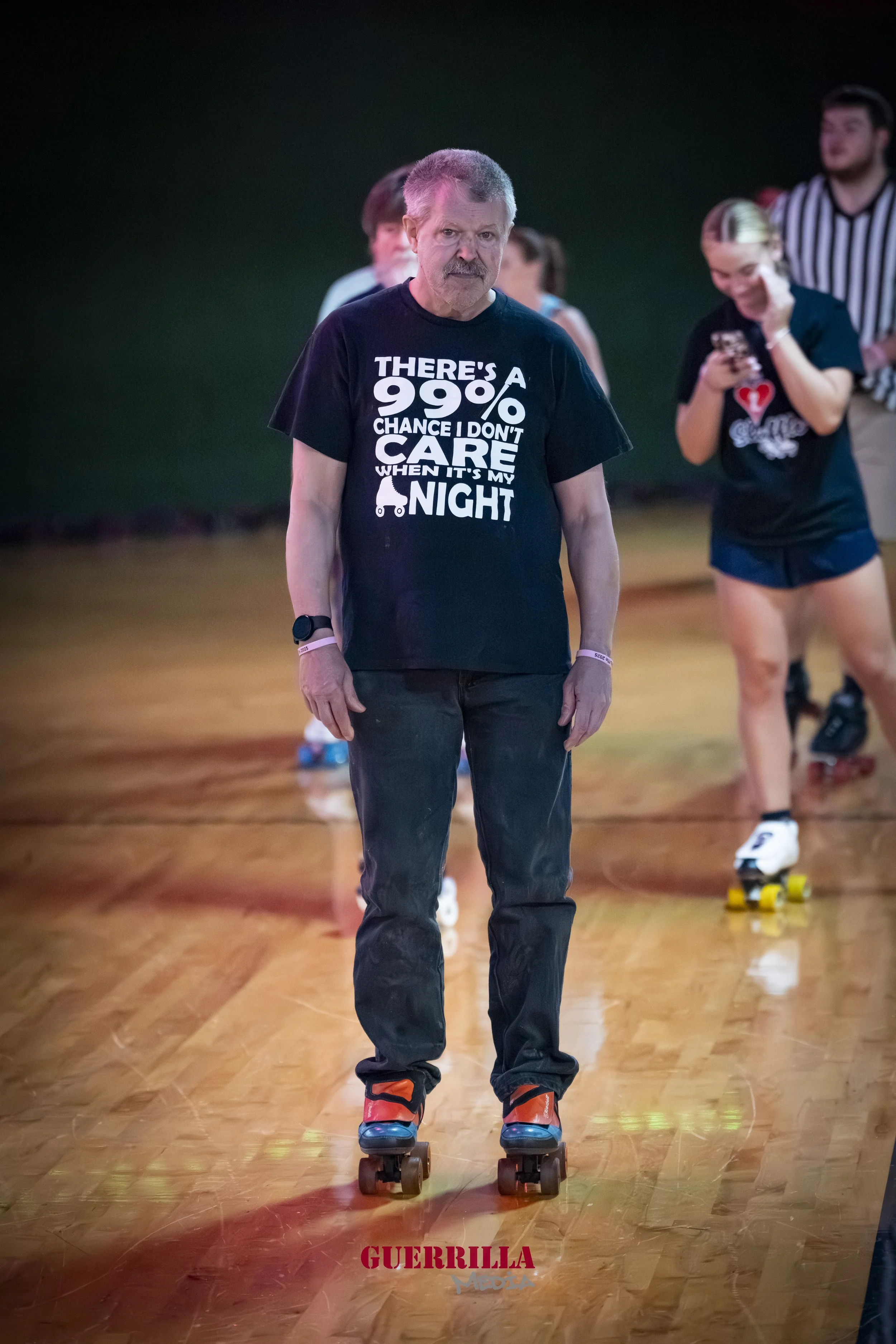 A man roller skating on a wooden floor, wearing a black T-shirt with white text, with people skating and socializing in the background.